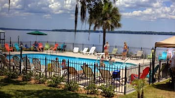 A swimming pool with lounge chairs and umbrellas, surrounded by a fence, with a view of a lake and trees in the background.