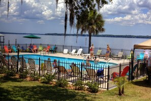 A swimming pool with lounge chairs and umbrellas, surrounded by a fence, with a view of a lake and trees in the background. - Crystal Cove Hotel (Palatka)