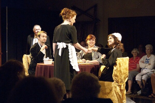 A stage with people seated at tables, some standing, and an audience in front.