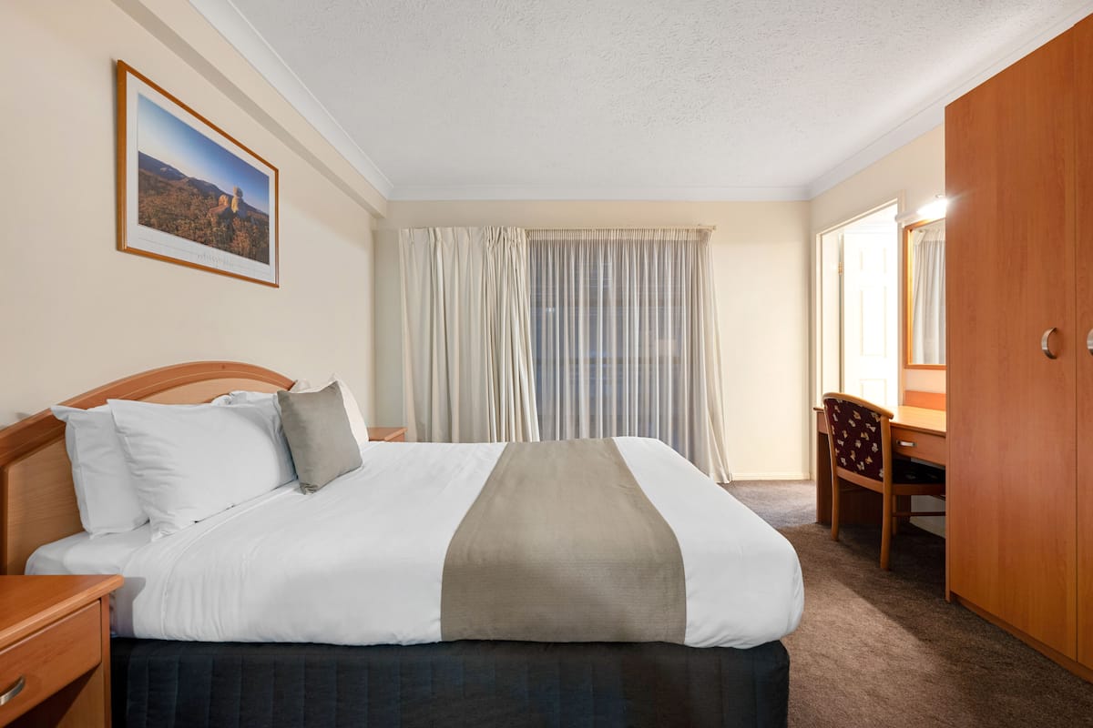 A hotel room with a bed, a desk, a chair, a wardrobe, and a large window with curtains.