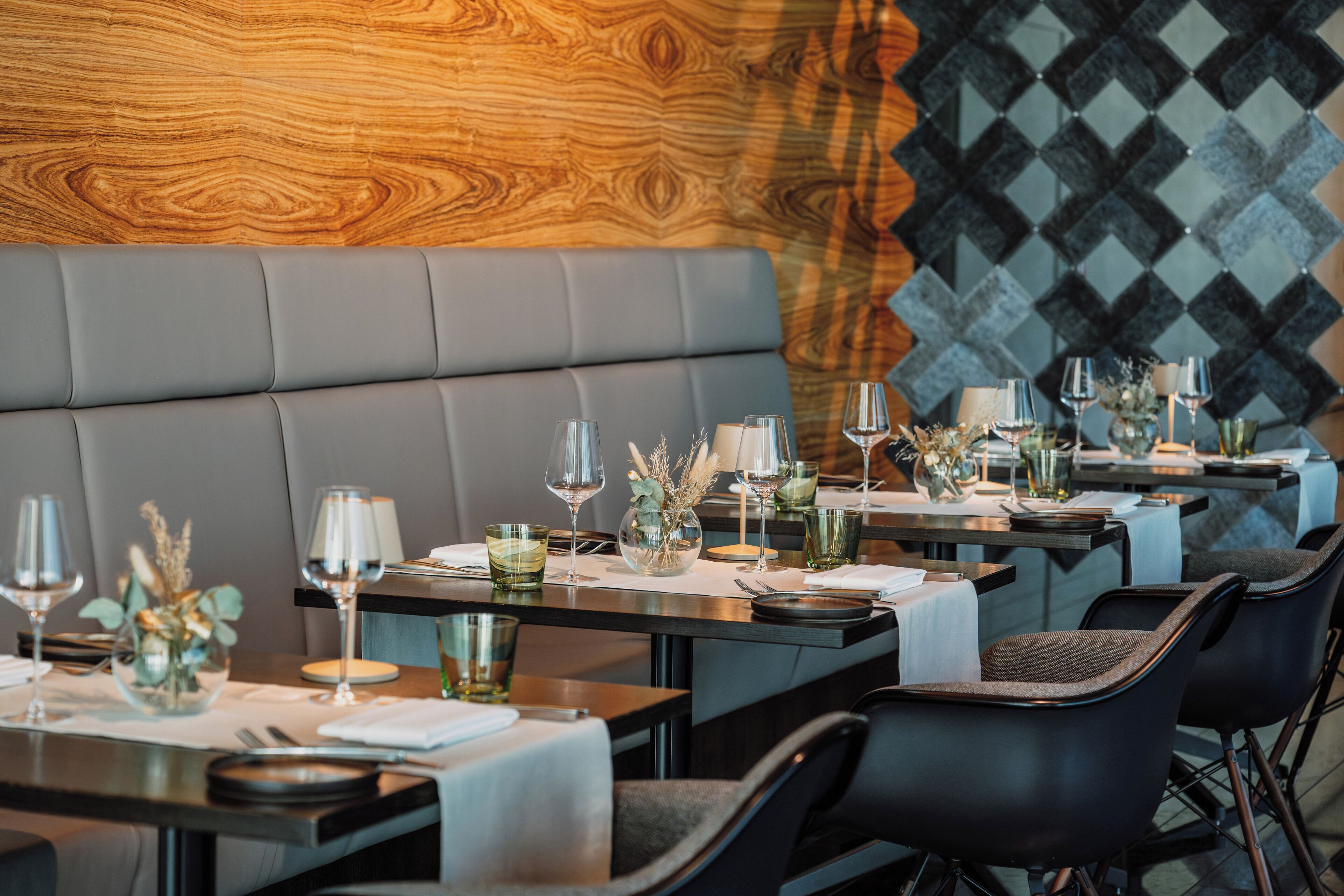 Ein Restaurant mit modernem Interieur, das Ledersitzbänke und eine Wand mit Muster bietet. Die Tische sind mit Weingläsern, Besteck und dekorativen Elementen gedeckt.