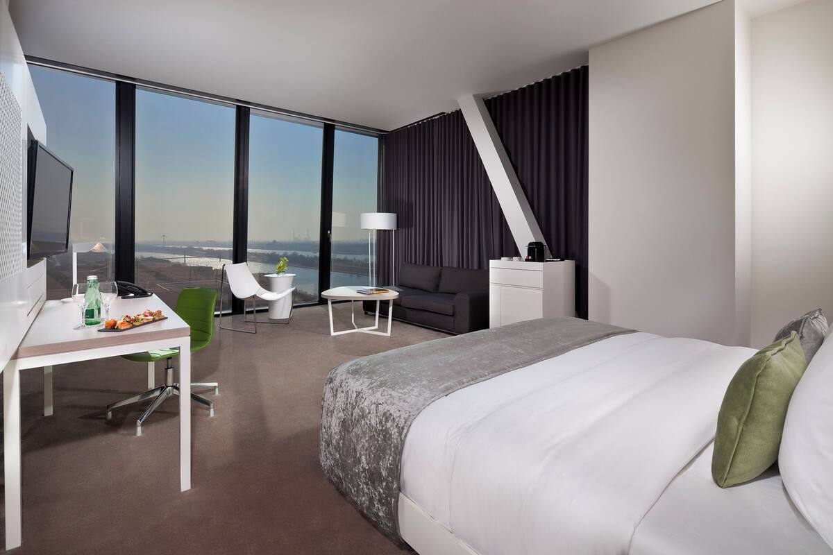 A modern hotel room with a large bed, a desk with a chair, a small table, and a sofa.