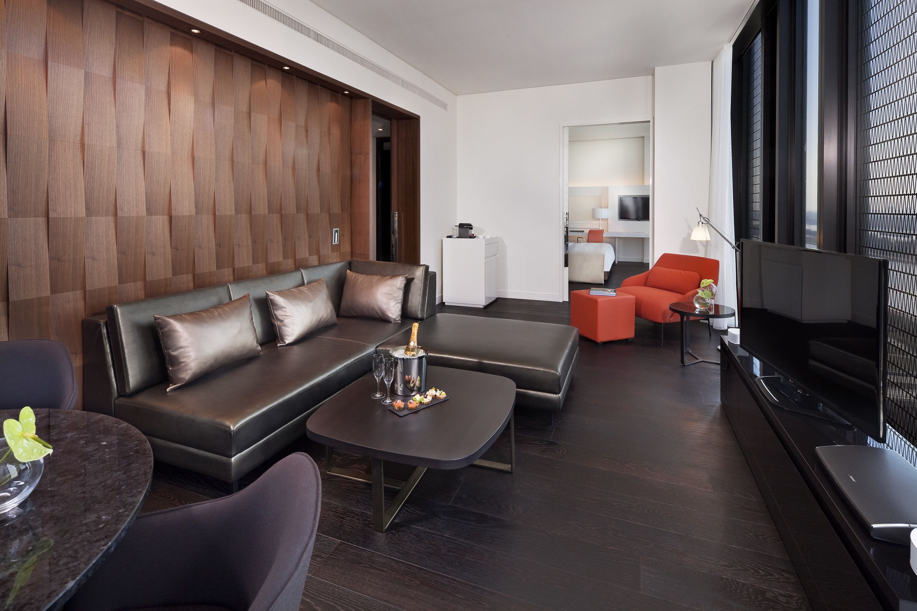 Una habitación de hotel moderna con un revestimiento de pared de madera oscura, un sofá de cuero, una pequeña mesa de comedor y un televisor de pantalla plana.