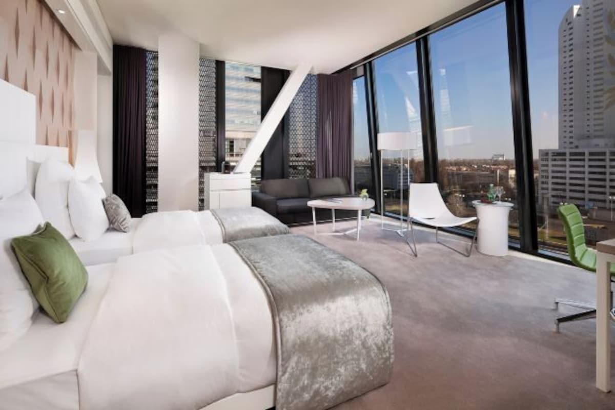 Una habitación de hotel moderna con una cama grande, un sofá, una mesita y vistas a la ciudad a través de amplios ventanales.