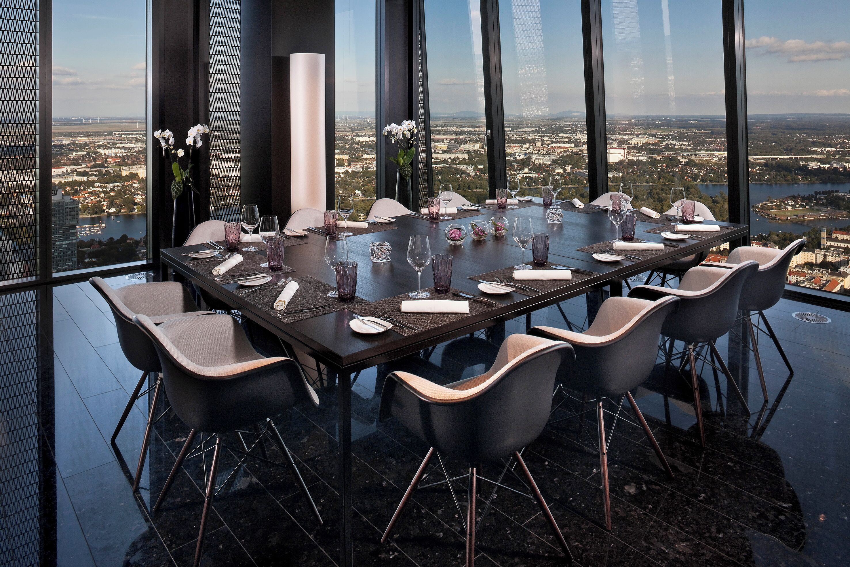A dining table set for eight with a panoramic city view, modern chairs, and elegant tableware.