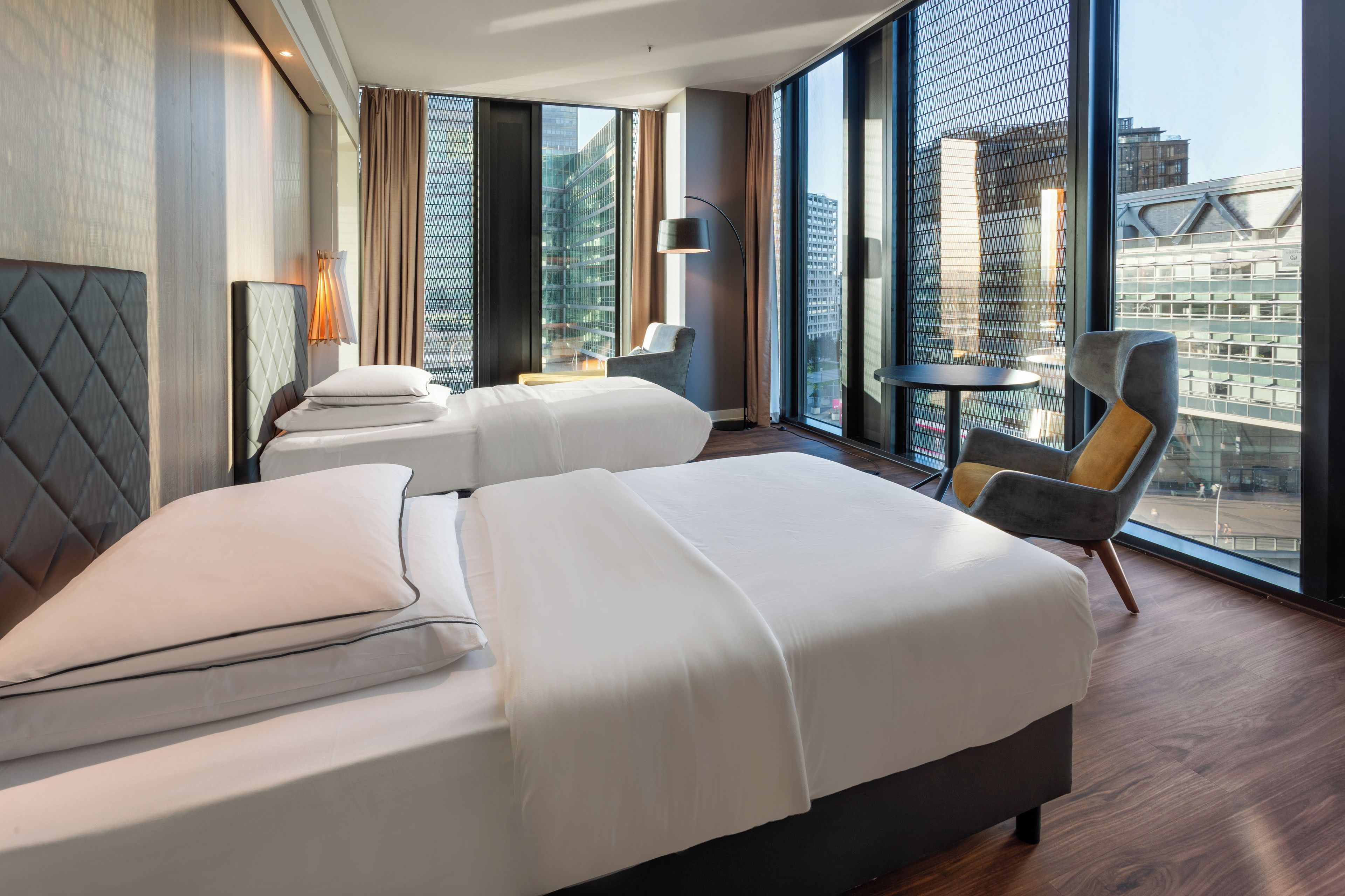 A hotel room with two beds, a small table, a chair, and large windows offering a city view.
