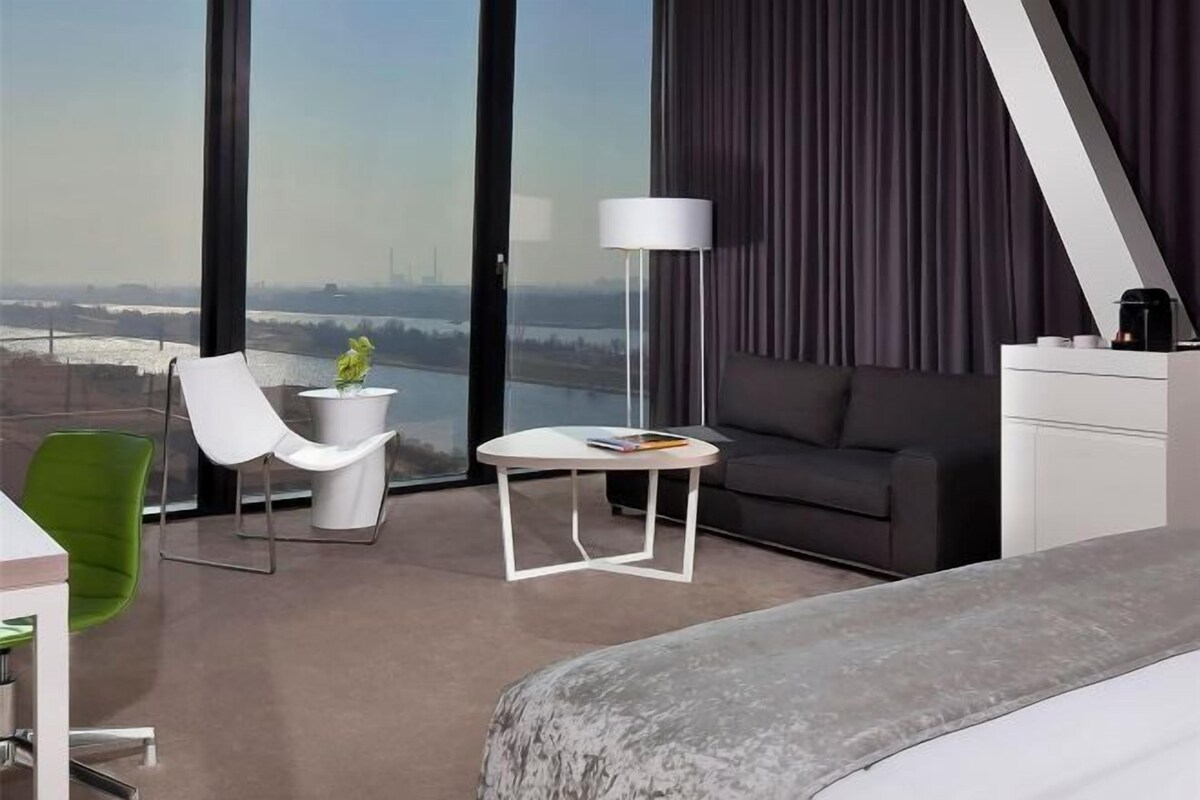 Habitación de hotel moderna con una cama grande, un sofá, una mesita y una silla. Se ve una masa de agua y un paisaje urbano a través de amplios ventanales.