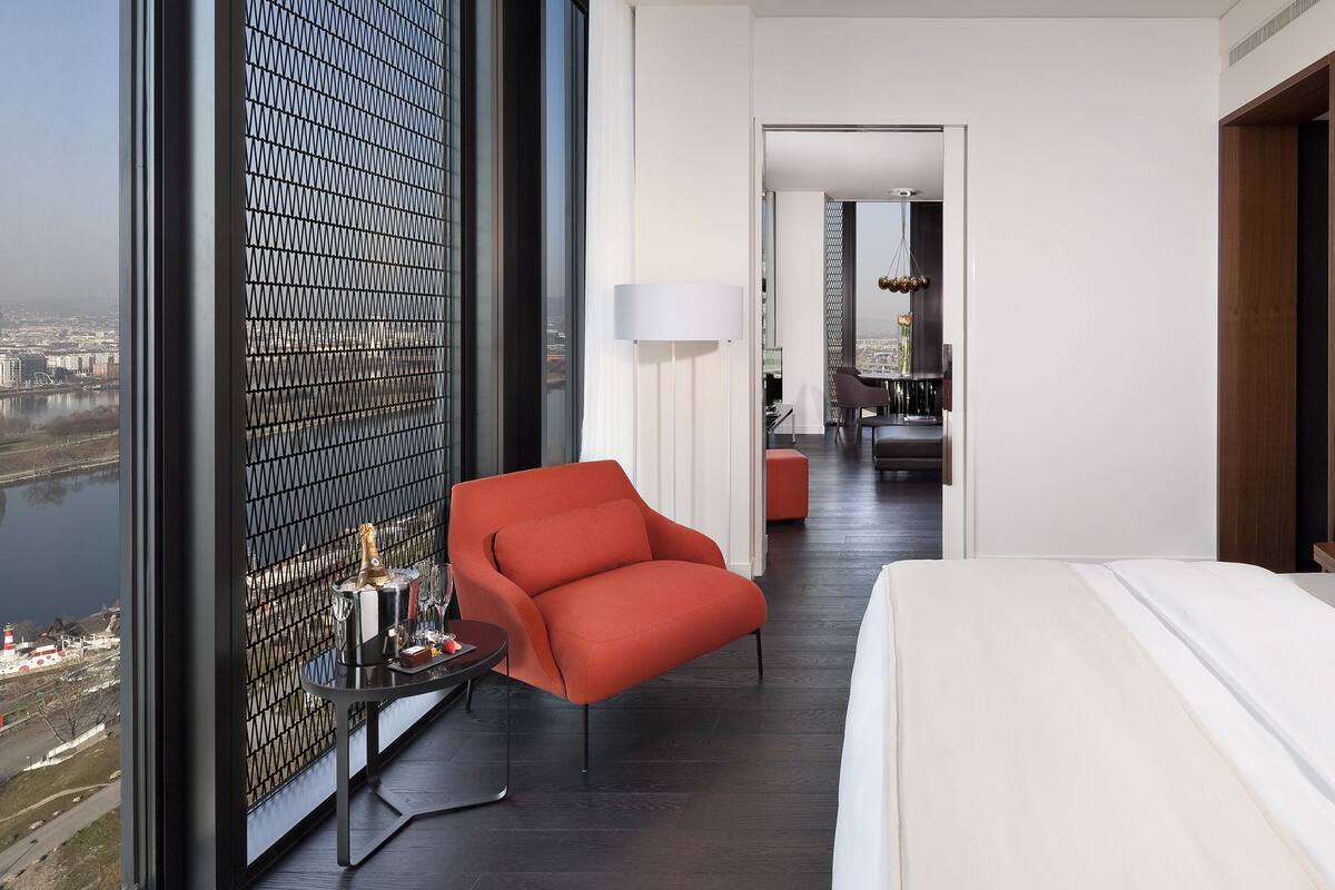 A modern hotel room with a large bed, a red armchair, a small table with a lamp, and a view of a cityscape through a window.