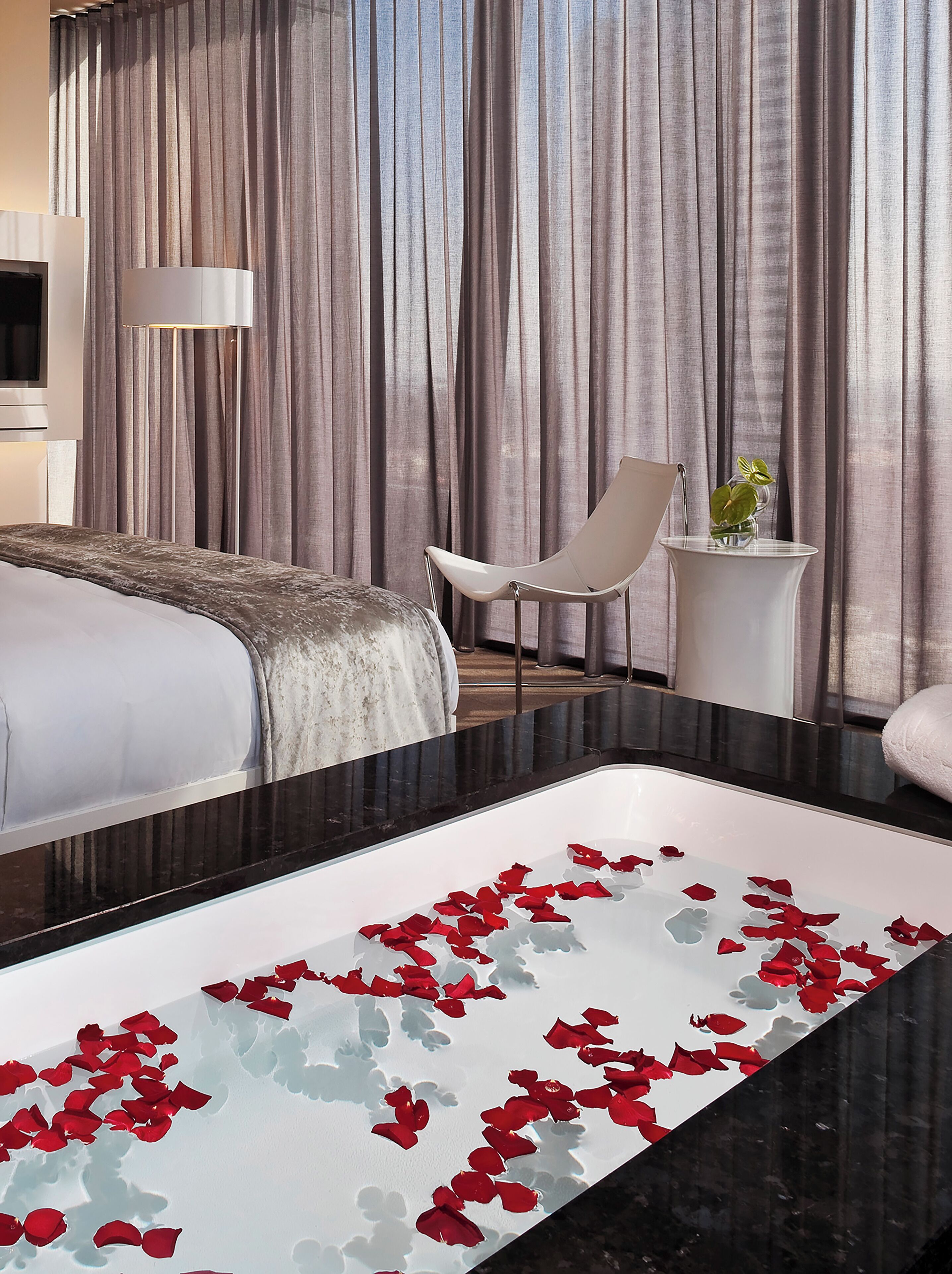 Habitación de hotel moderna con una cama grande, una silla blanca, una mesita con una planta y una mesa de vidrio con pétalos de rosa.