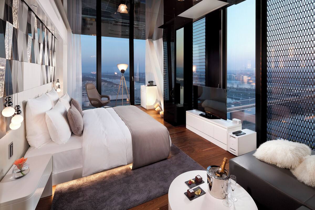 A modern bedroom with a large bed, bedside tables, a TV, and a city view.