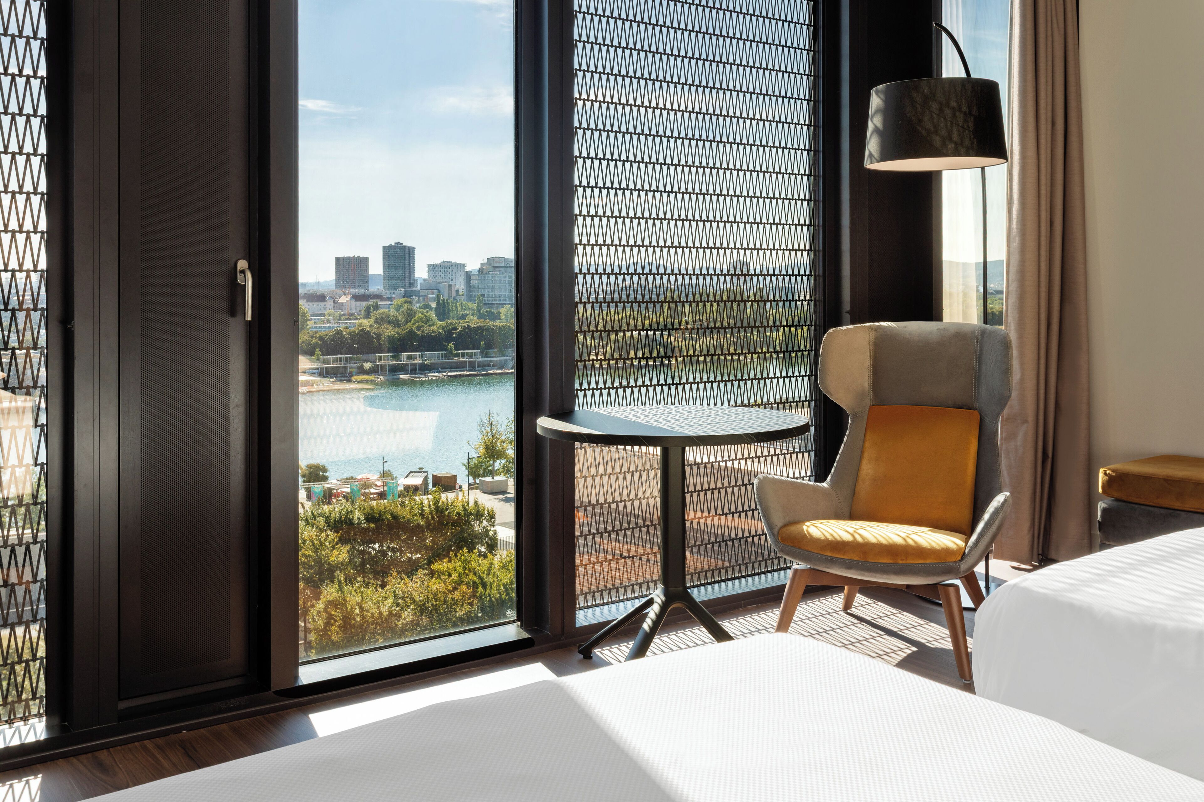 Een hotelkamer met een groot raam met uitzicht over de stad, een klein rond tafeltje, een moderne stoel, een bed en een vloerlamp.