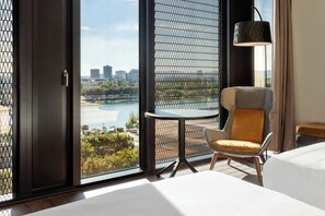 A hotel room with a large window offering a city view, a small round table, a modern chair, a bed, and a floor lamp.