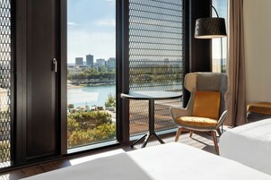 A hotel room with a large window offering a city view, a small round table, a modern chair, a bed, and a floor lamp.