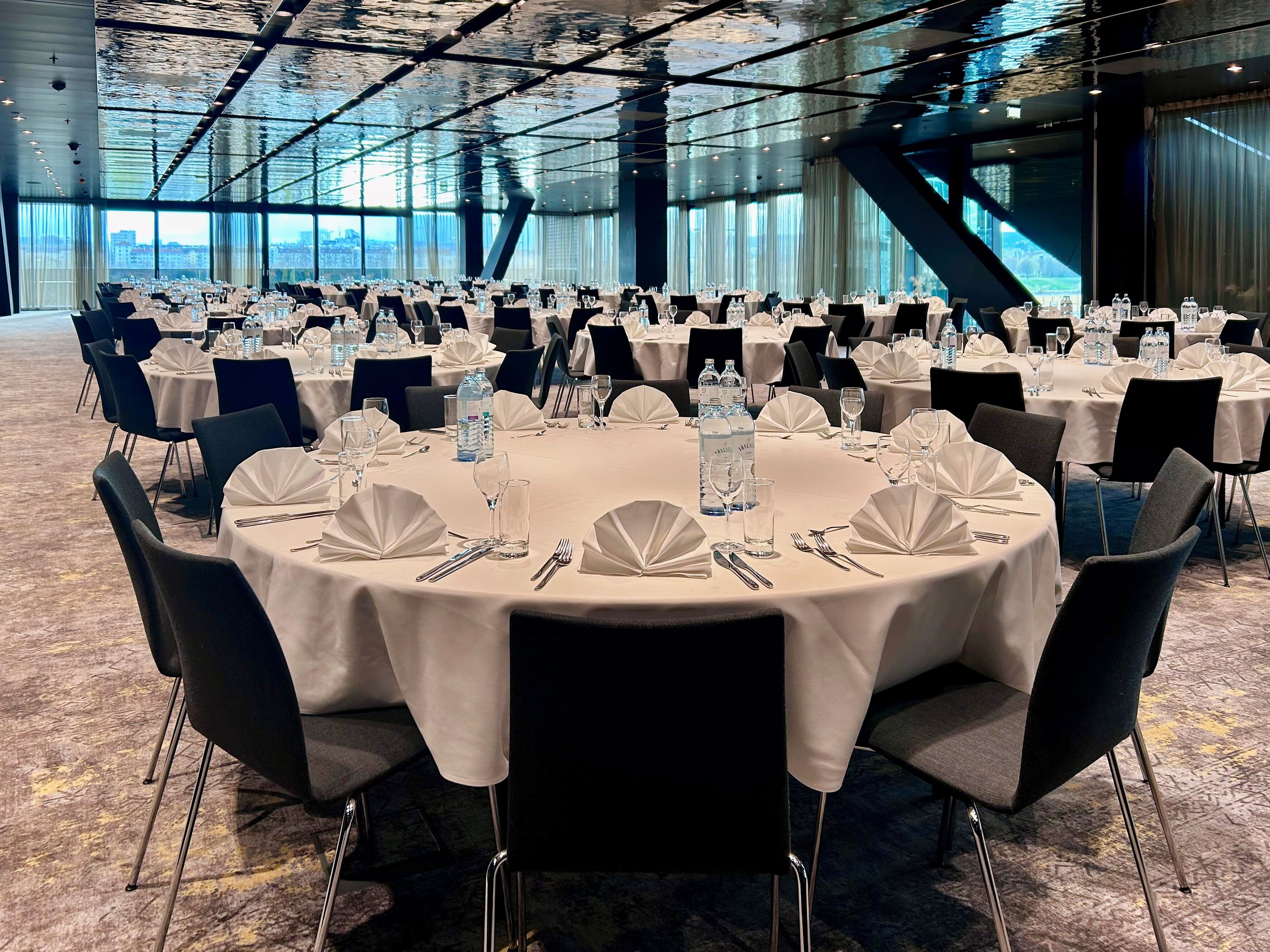 Een ruime balzaal met ronde tafels gedekt voor een formeel evenement, met witte tafelkleden, zwarte stoelen en waterflesjes.