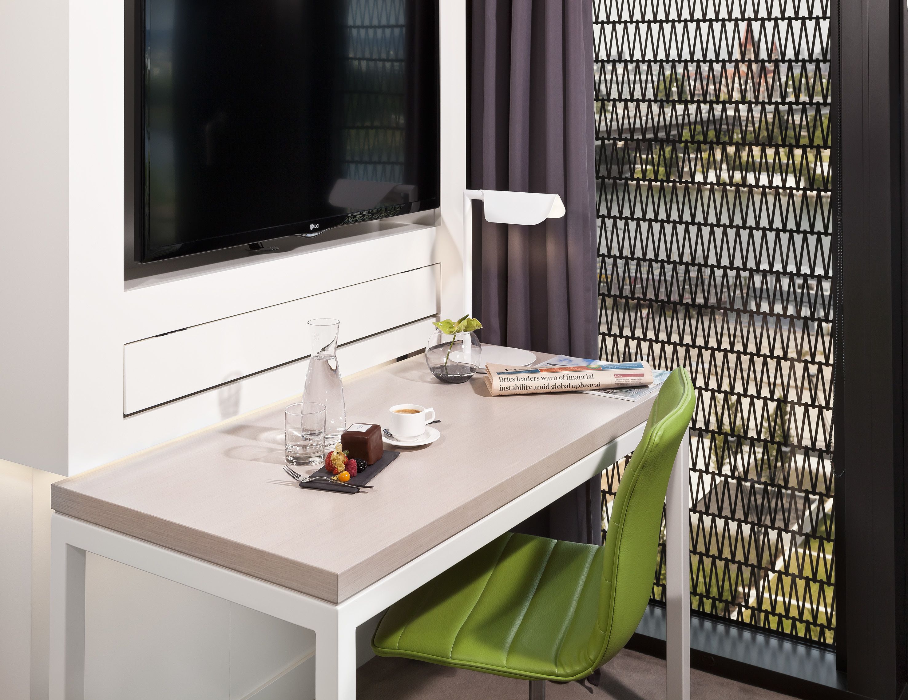 A modern hotel room with a desk, a TV, a water pitcher, a cup of coffee, a magazine, and a green chair.