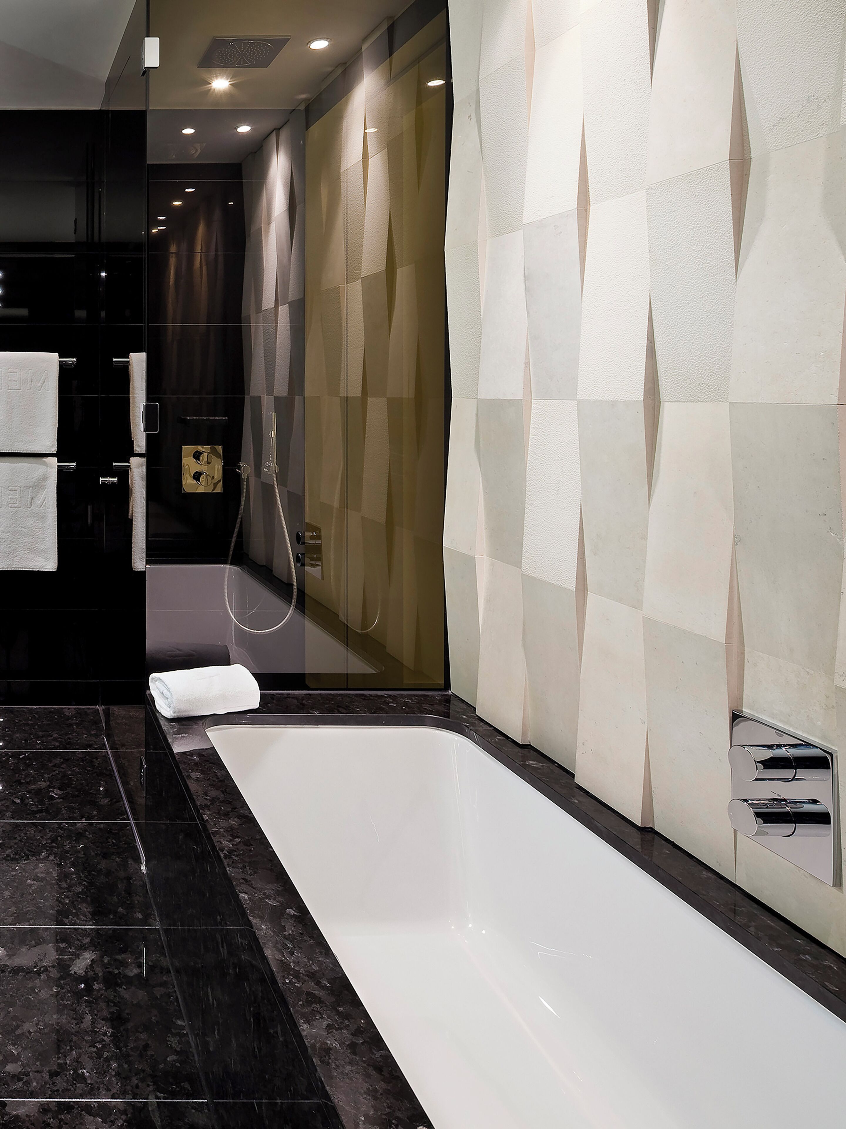 Un baño moderno con una bañera grande, cabina de ducha de vidrio y pared revestida con un diseño geométrico.