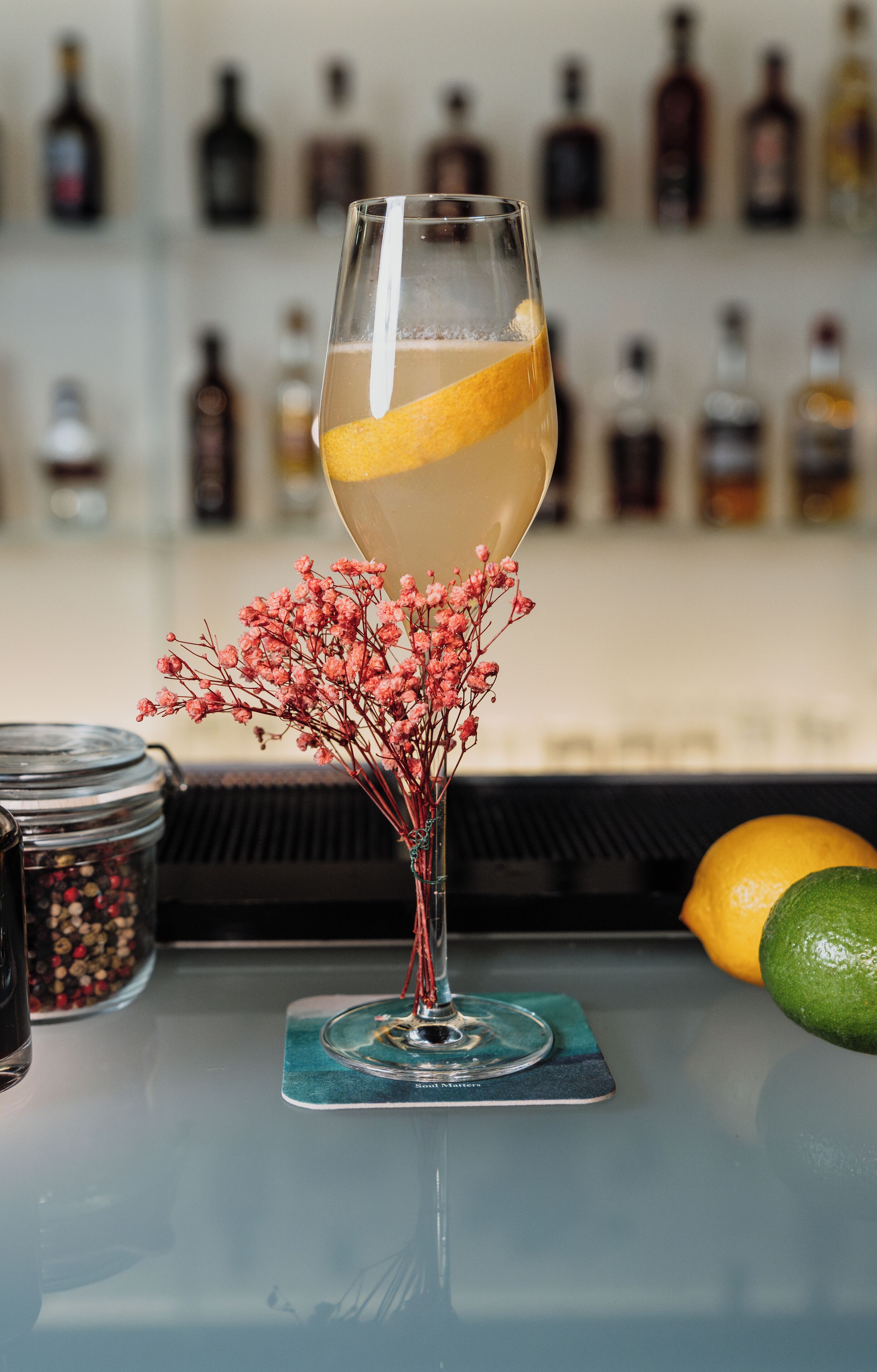 Ein Cocktailglas mit Zitronenscheibe auf einem Untersetzer, ein Gewürzglas und eine Vase mit getrockneten Blumen auf einer Bartheke.