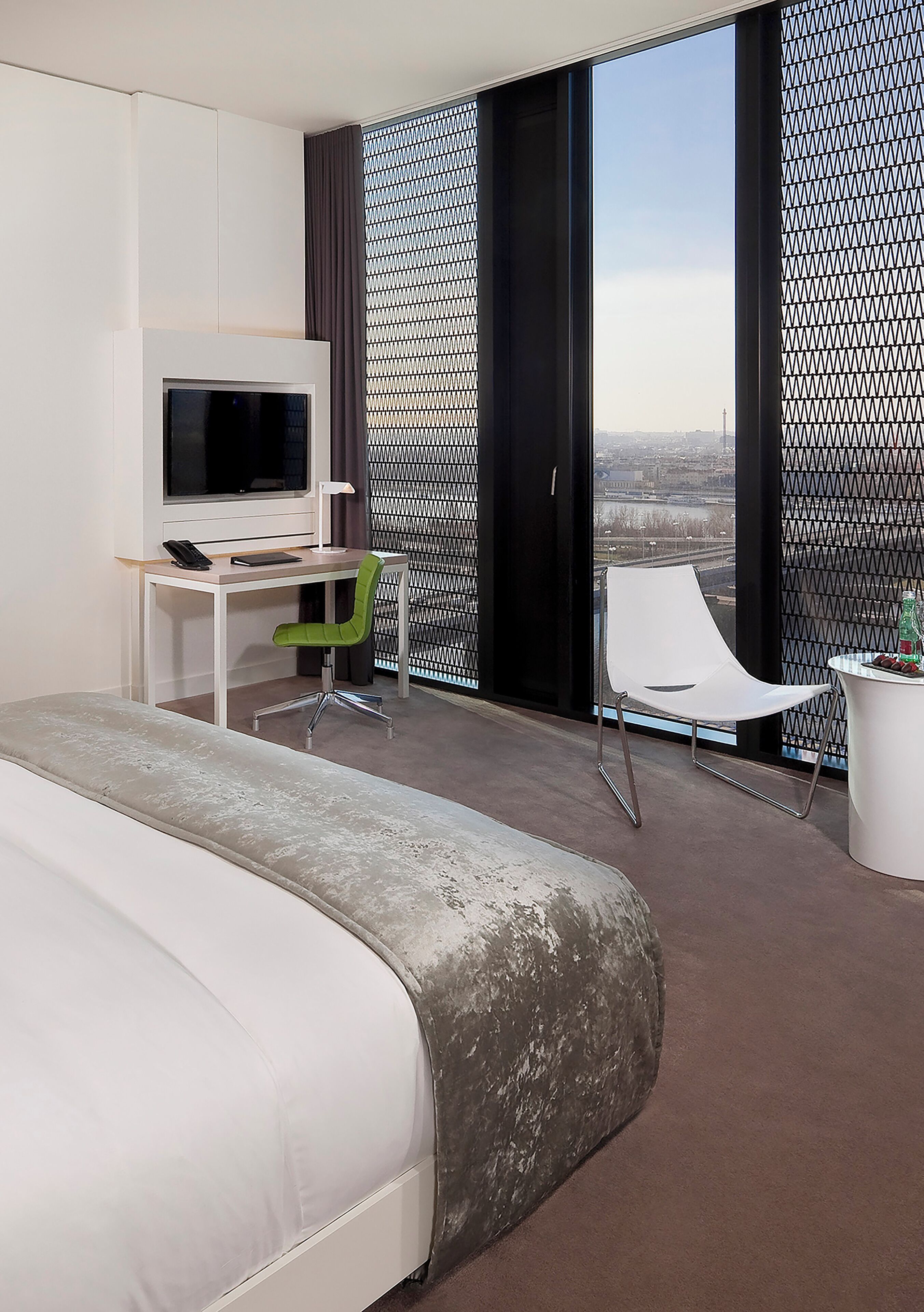 A modern hotel room with a large bed, a desk with a TV, a green chair, and a balcony with a city view.