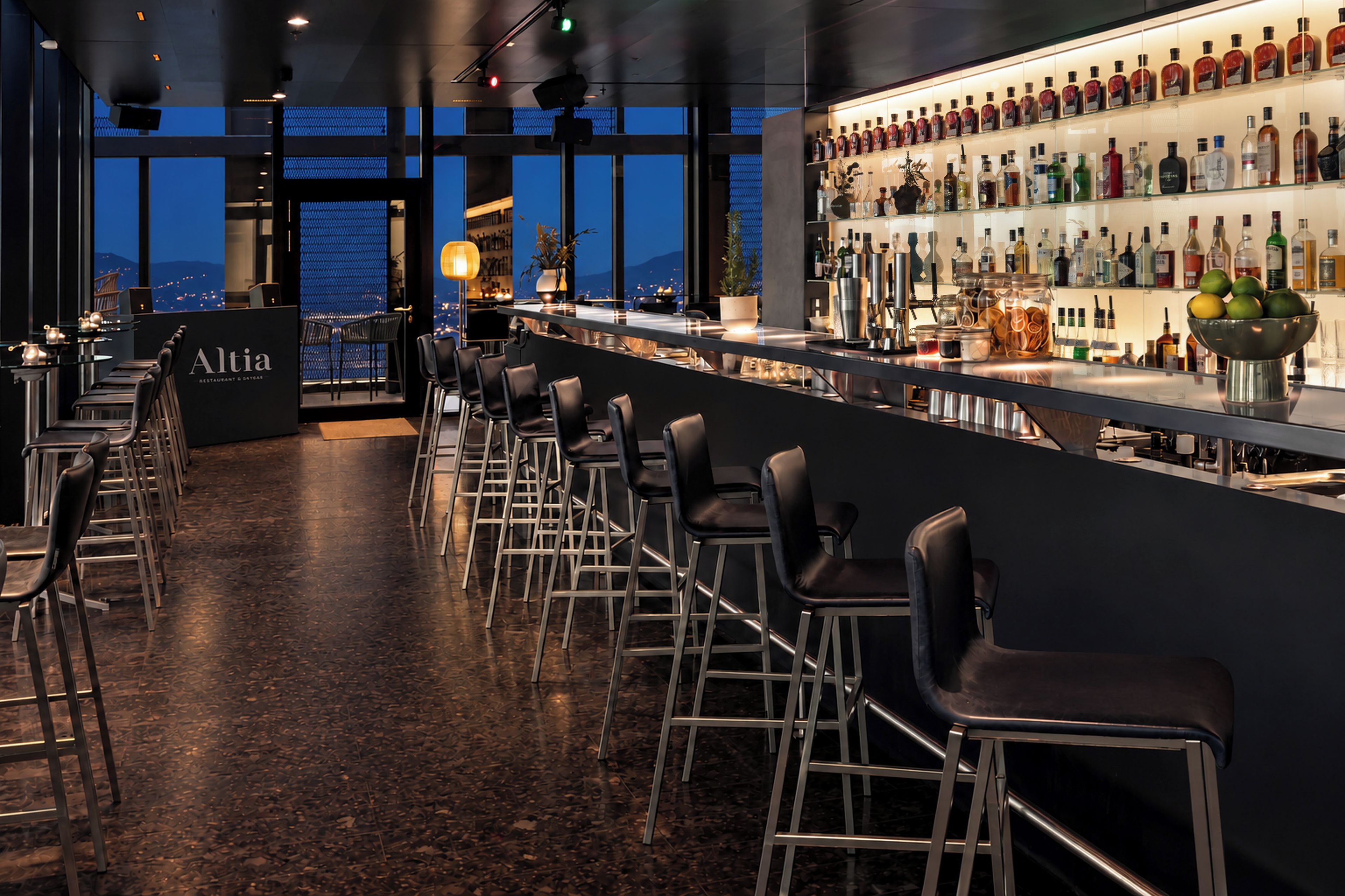 A bar area with a long counter, multiple stools, and a variety of bottles on shelves.