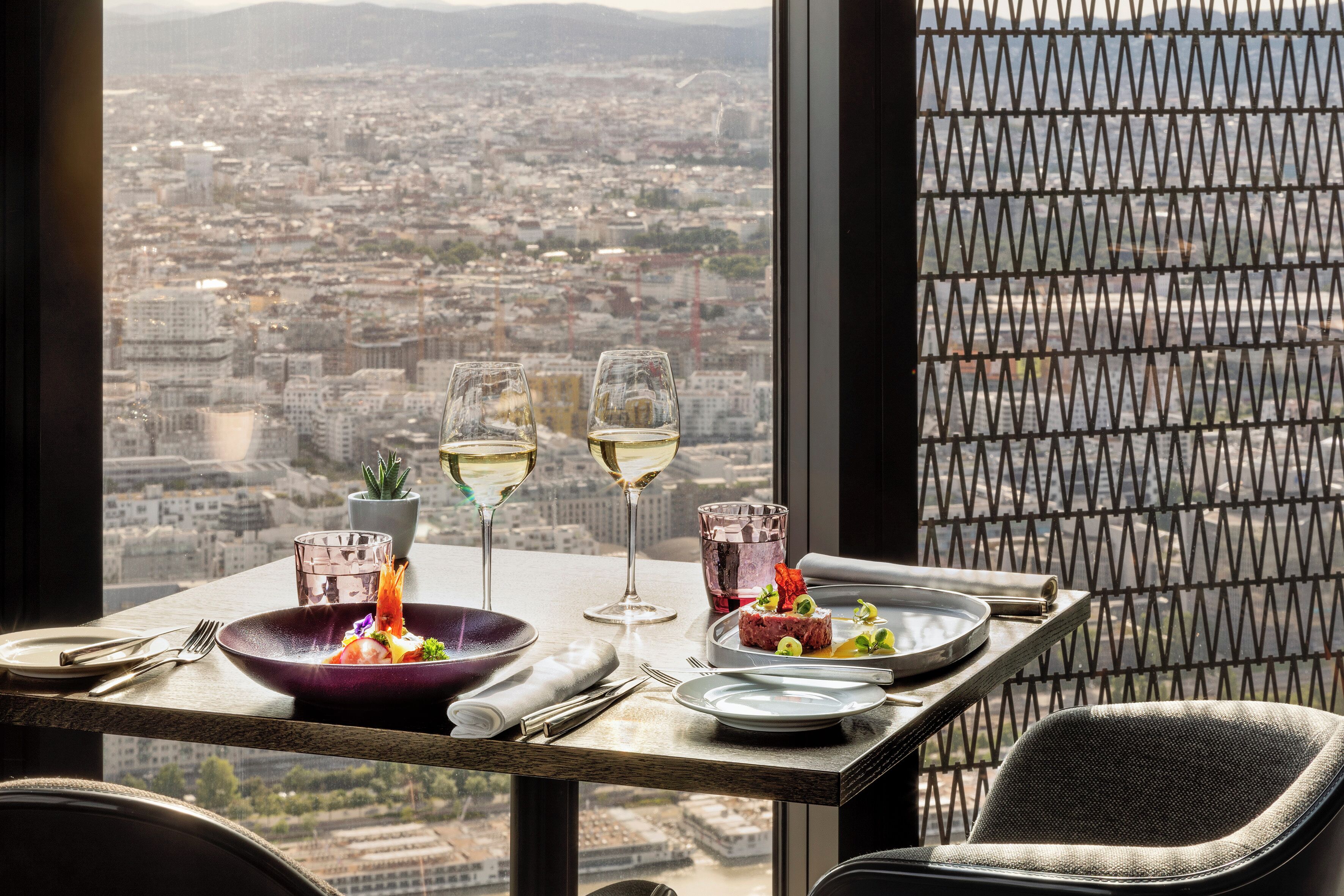 Une table avec vue sur la ville, deux verres de vin blanc, un bol violet et une assiette de nourriture.