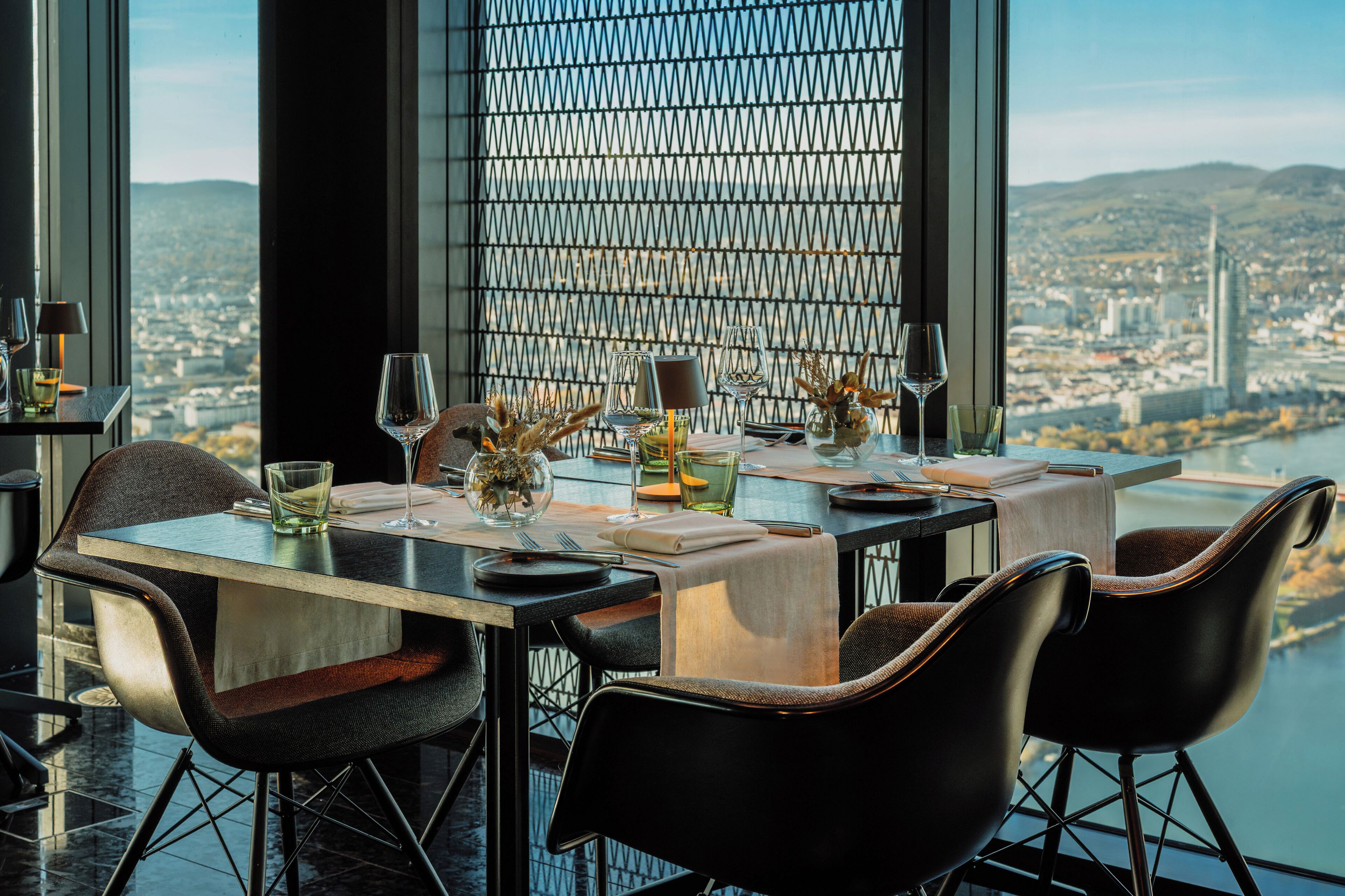 Zona pranzo con tavolo apparecchiato per due, vista sulla città attraverso una finestra e composizione floreale decorativa al centro.