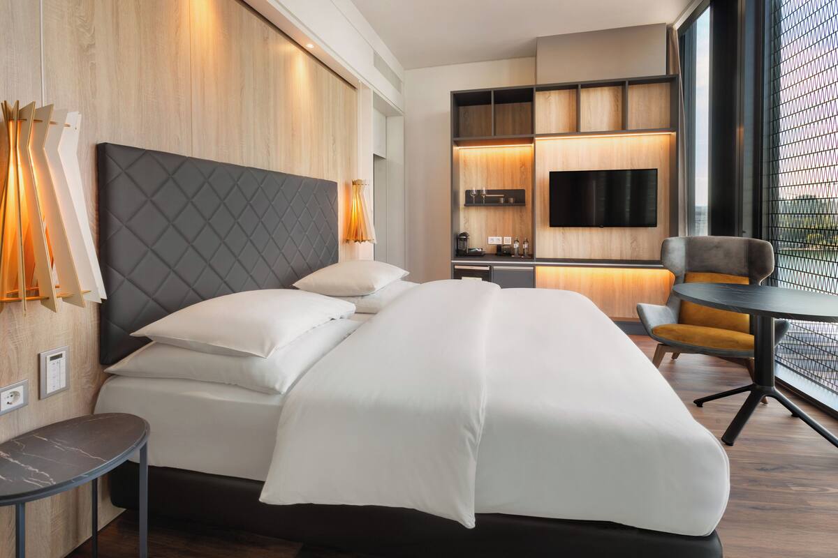 A modern hotel room with a large bed, a small table, a seating area, and a TV mounted on the wall.