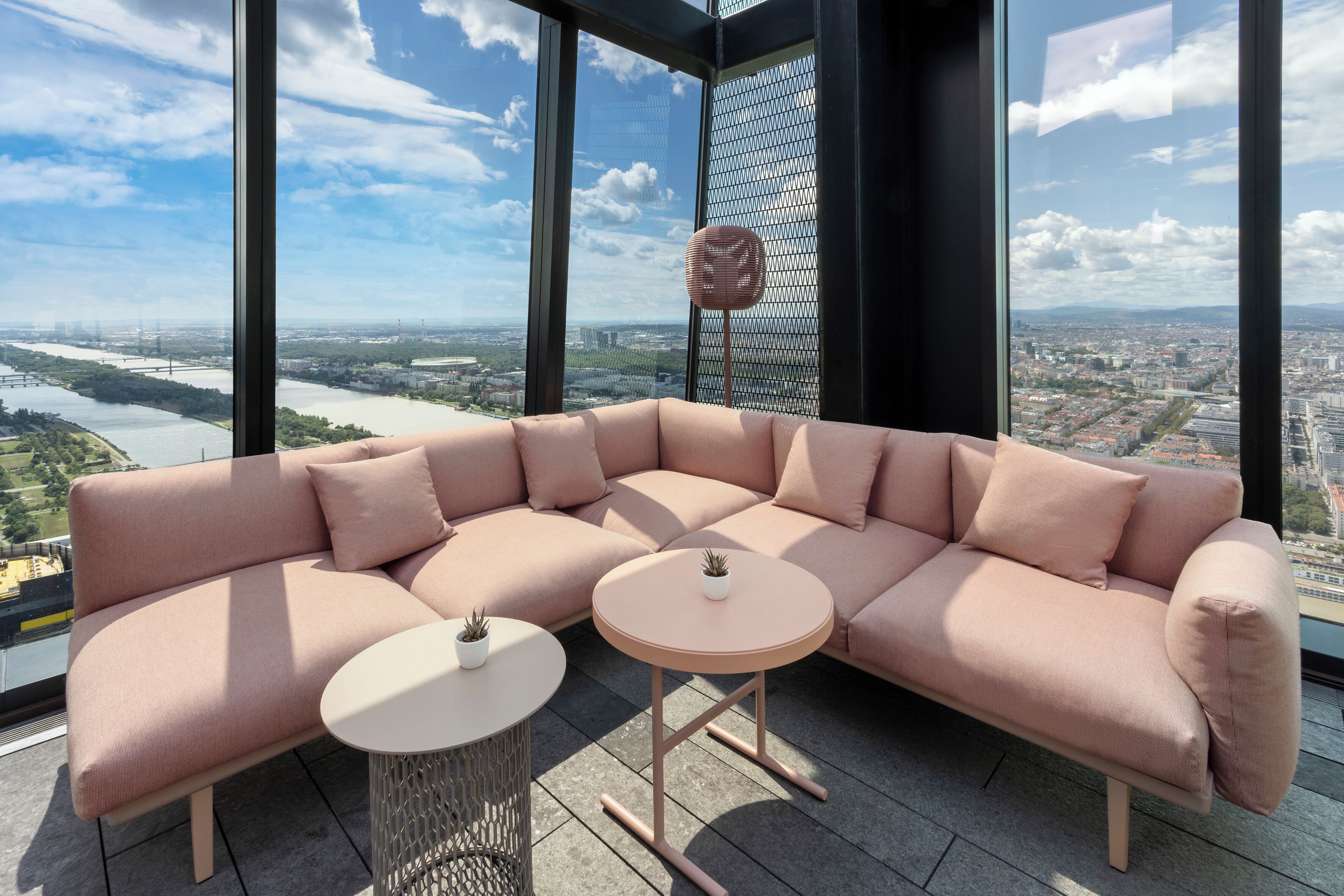 Ein modernes, pinkes Modulsofa mit zwei runden Tischen, einer mit einer kleinen Pflanze. Große Fenster bieten Blick auf eine Stadtlandschaft und einen Fluss.