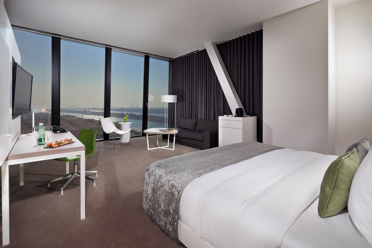 Quarto de hotel moderno com uma cama grande, uma escrivaninha com cadeira, uma mesinha e um sofá.