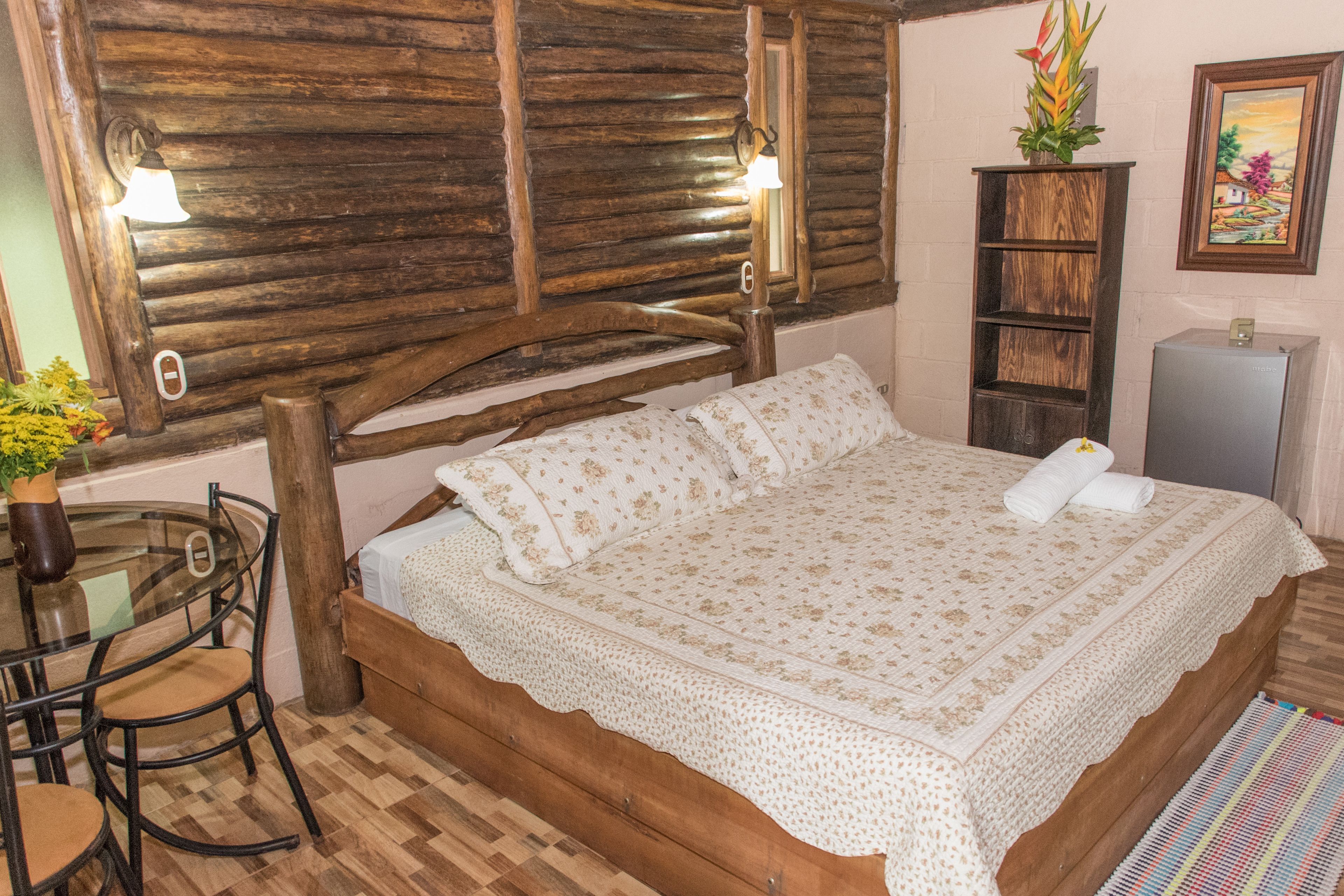 A wooden bed with a floral bedspread, a small table with chairs, and a wall-mounted shelf with a plant.