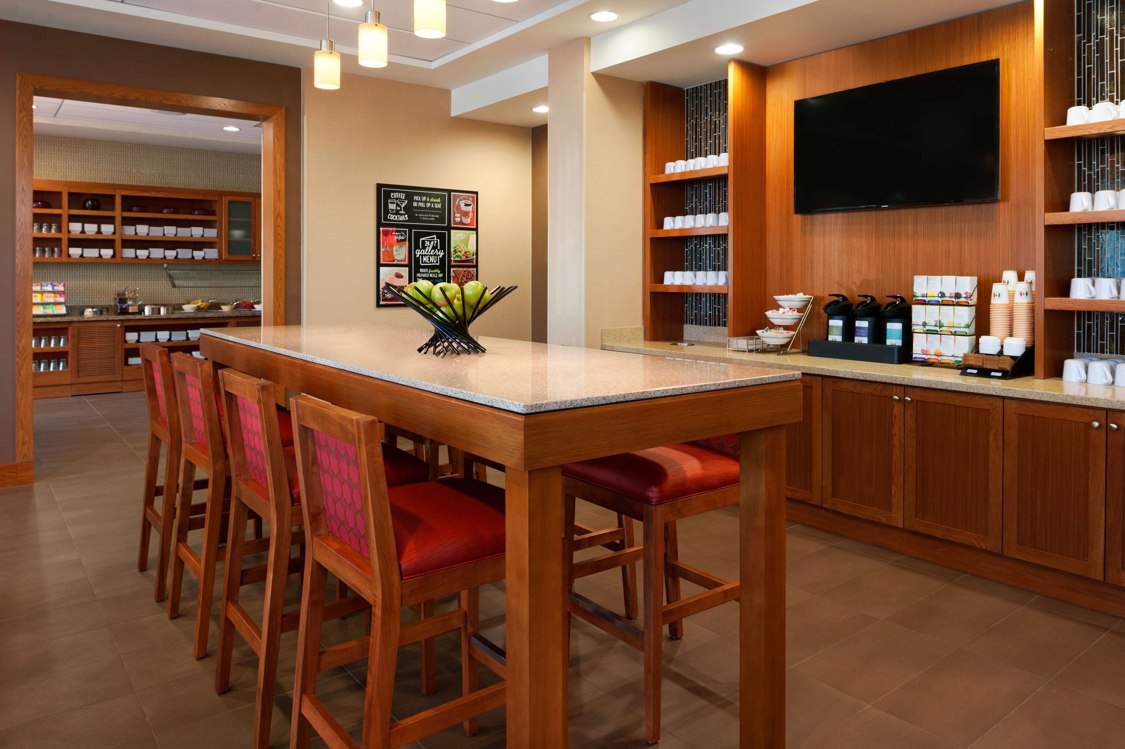 Un vestíbulo de hotel con un mostrador de recepción de madera, sillas con cojines rojos, una estación de café y un televisor montado en la pared.