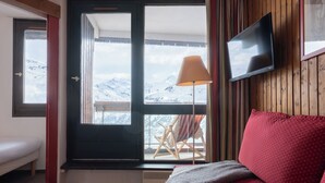 A cozy room with a wooden wall, a TV mounted above a red sofa, a window with a view of snow-capped mountains, and a chair by the window. - Résidence Pierre & Vacances Les Menuires Les Combes (Les Belleville)