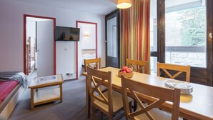 A room with a bed, a TV mounted on the wall, a dining table with chairs, and a window with a view of trees. - Résidence Pierre & Vacances Les Menuires Les Combes (Les Belleville)