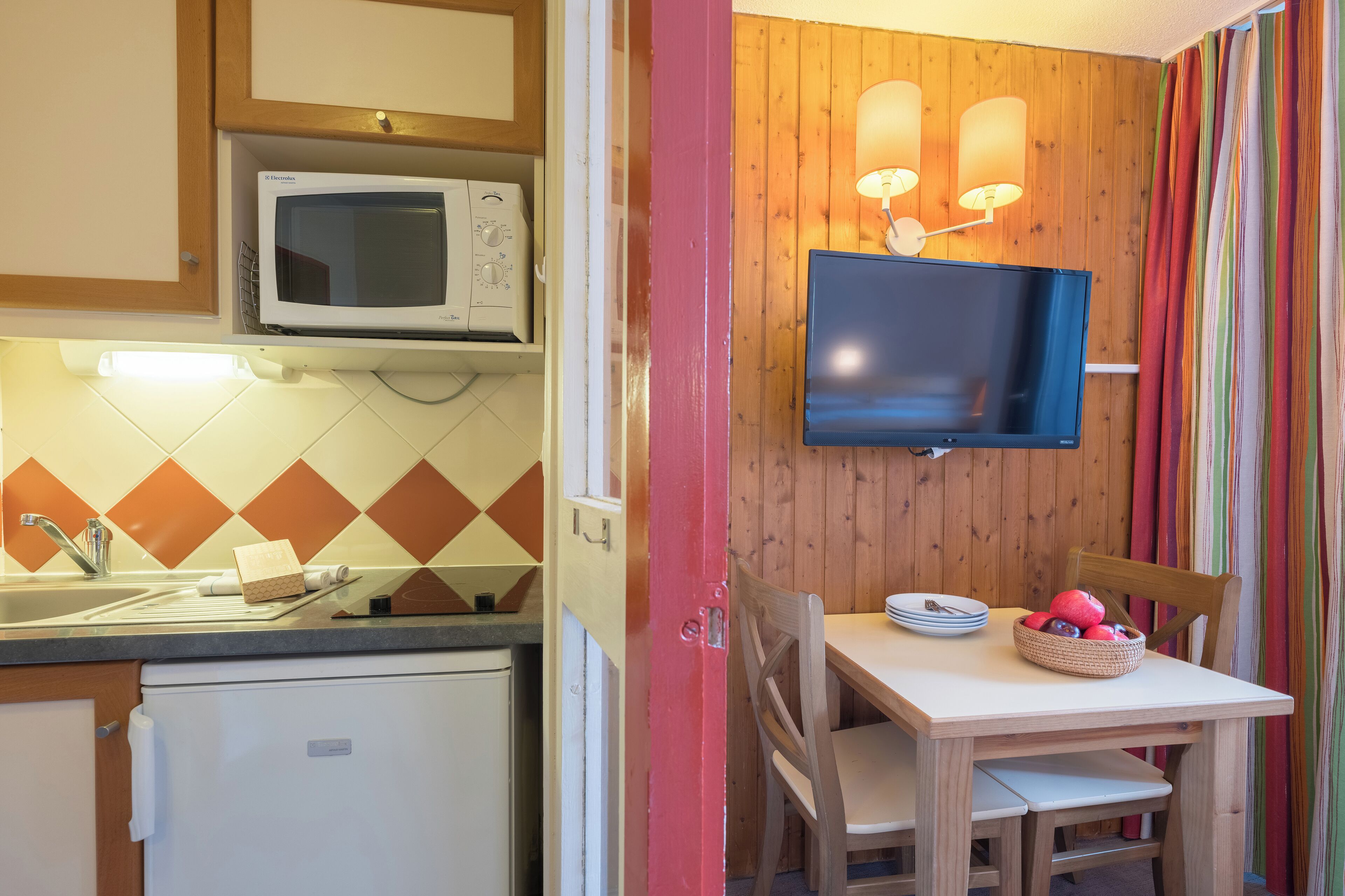 Une cuisine compacte équipée d’un four à micro-ondes, d’un petit réfrigérateur, d’une table à manger avec des chaises et d’un téléviseur à écran plat fixé au mur.