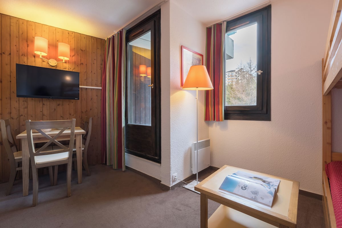 Ett hotellrum med en väggpanel av trä, en tv som hänger ovanför ett träbord, ett fönster med gardiner, en lampa och en säng med ett rött överkast.
