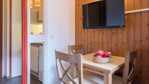 A dining area with a wooden wall, a flat-screen TV mounted above a table, and a basket of fruit. - Résidence Pierre & Vacances Les Menuires Les Combes (Les Belleville)