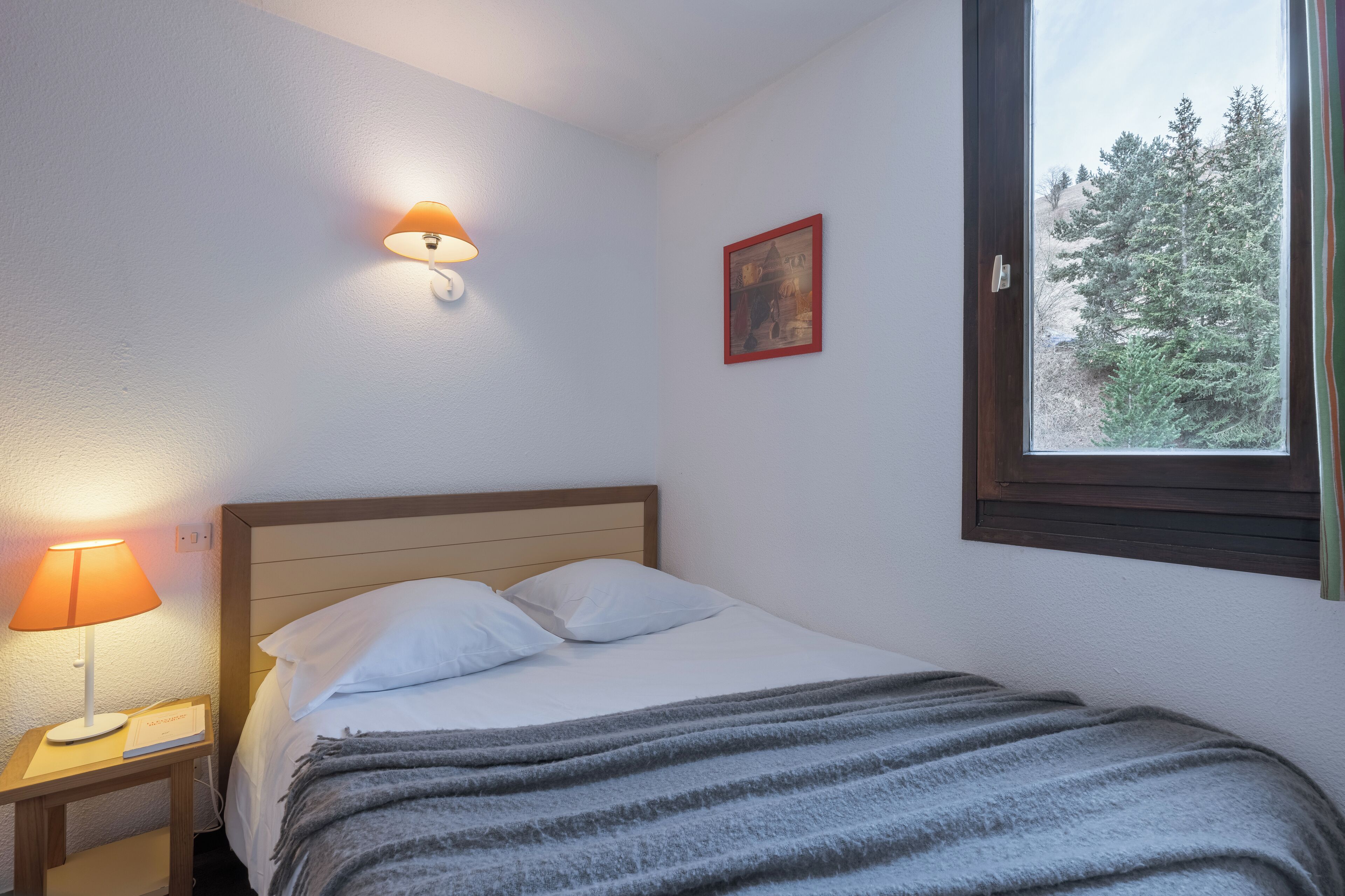 Ein ordentlich bezogenes Bett mit weißen Leinen und einer Streifen-Decke, eine Nachttischlampe, ein gerahmtes Bild an der Wand und ein Fenster mit Blick auf Bäume.