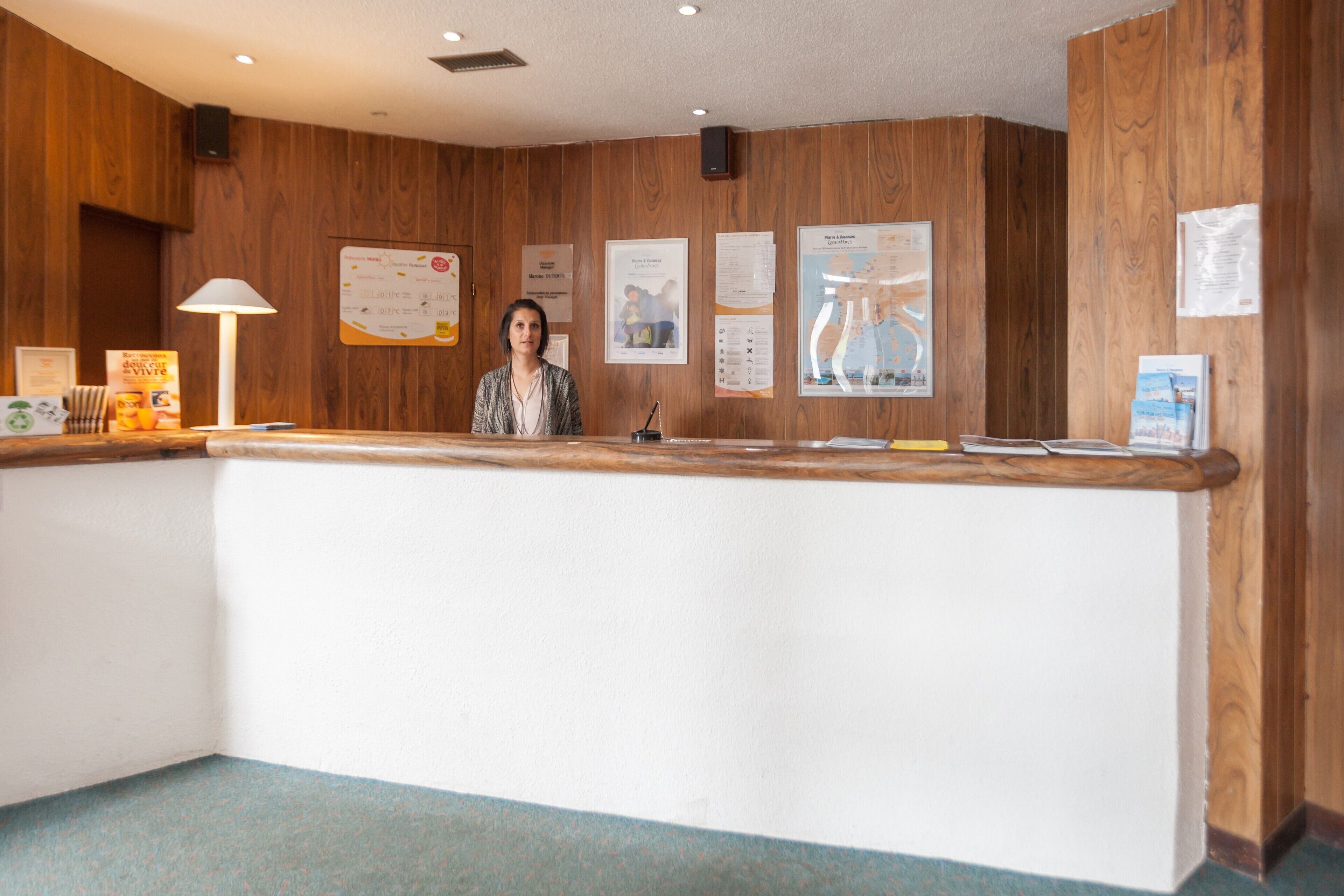 Un mostrador de recepción con una mujer de pie detrás, paneles de madera en las paredes y varios carteles informativos.