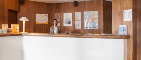Un mostrador de recepción con una mujer de pie detrás, paneles de madera en las paredes y varios carteles informativos.