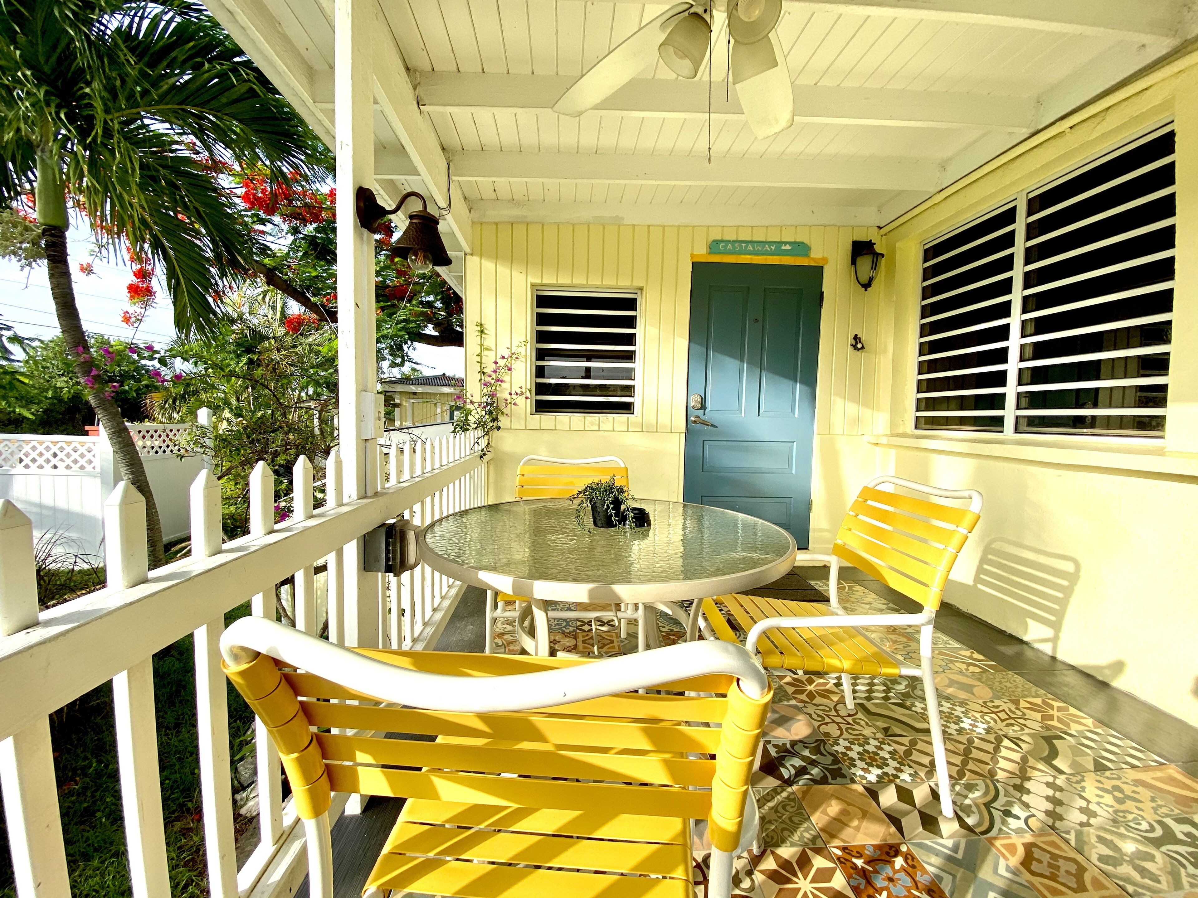 Une terrasse couverte avec une table ronde et des chaises jaunes, une porte bleue et une clôture blanche en bois.