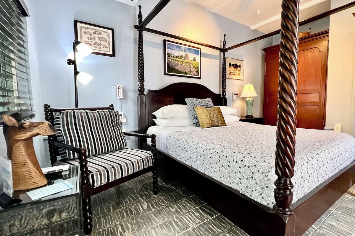 A four-poster bed with a patterned bedspread, a striped chair, and a wooden sculpture.