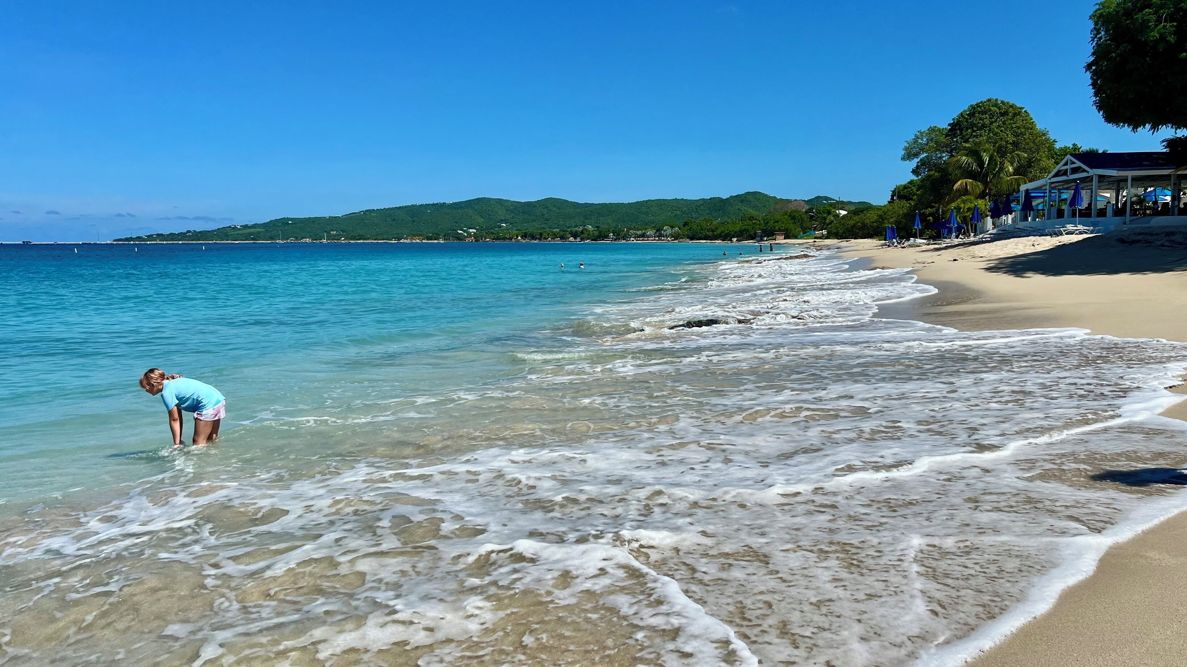 A beach with clear water, a person wading in the shallow part, and a covered area with umbrellas on the right.