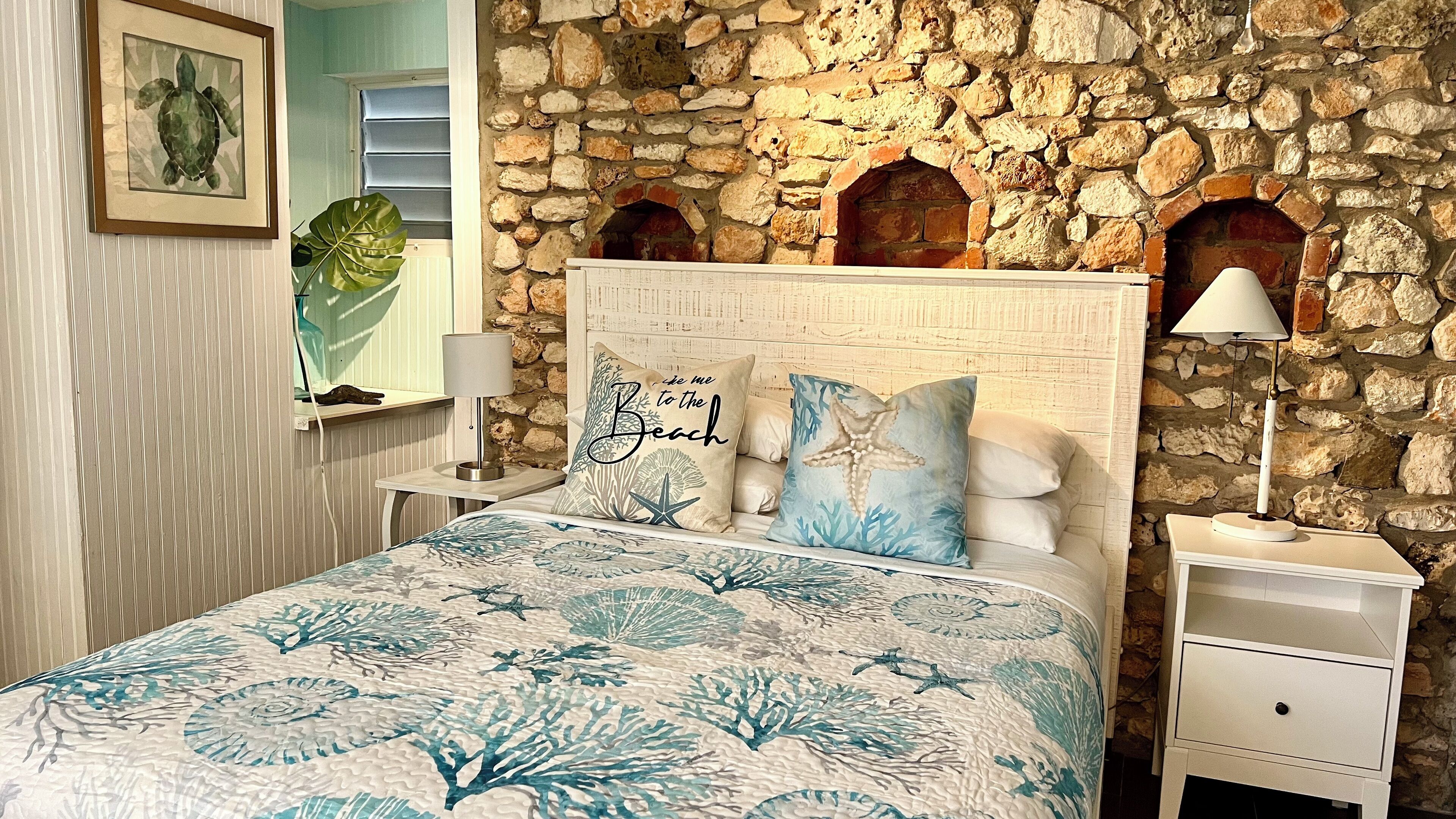 A bedroom with a stone wall, a bed with a beach-themed bedspread, a nightstand with a lamp, and a framed picture on the wall.