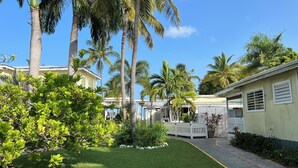 A residential area with palm trees, a white picket fence, and a pathway leading to a building with a window and a door. - Cottages by the Sea (Frederiksted)