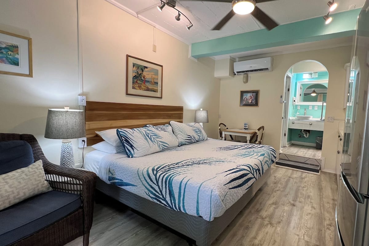 A hotel room with a bed, a chair, a desk, a bathroom visible through an archway, and an air conditioning unit on the wall.
