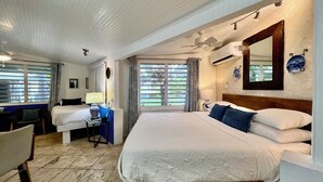 A hotel room with a large bed, a desk with a chair, a window with blinds, a ceiling fan, and an air conditioning unit. - Cottages by the Sea (Frederiksted)