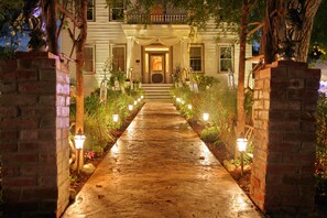 A well-lit pathway leading to a building with a front porch, surrounded by plants and decorative lights.