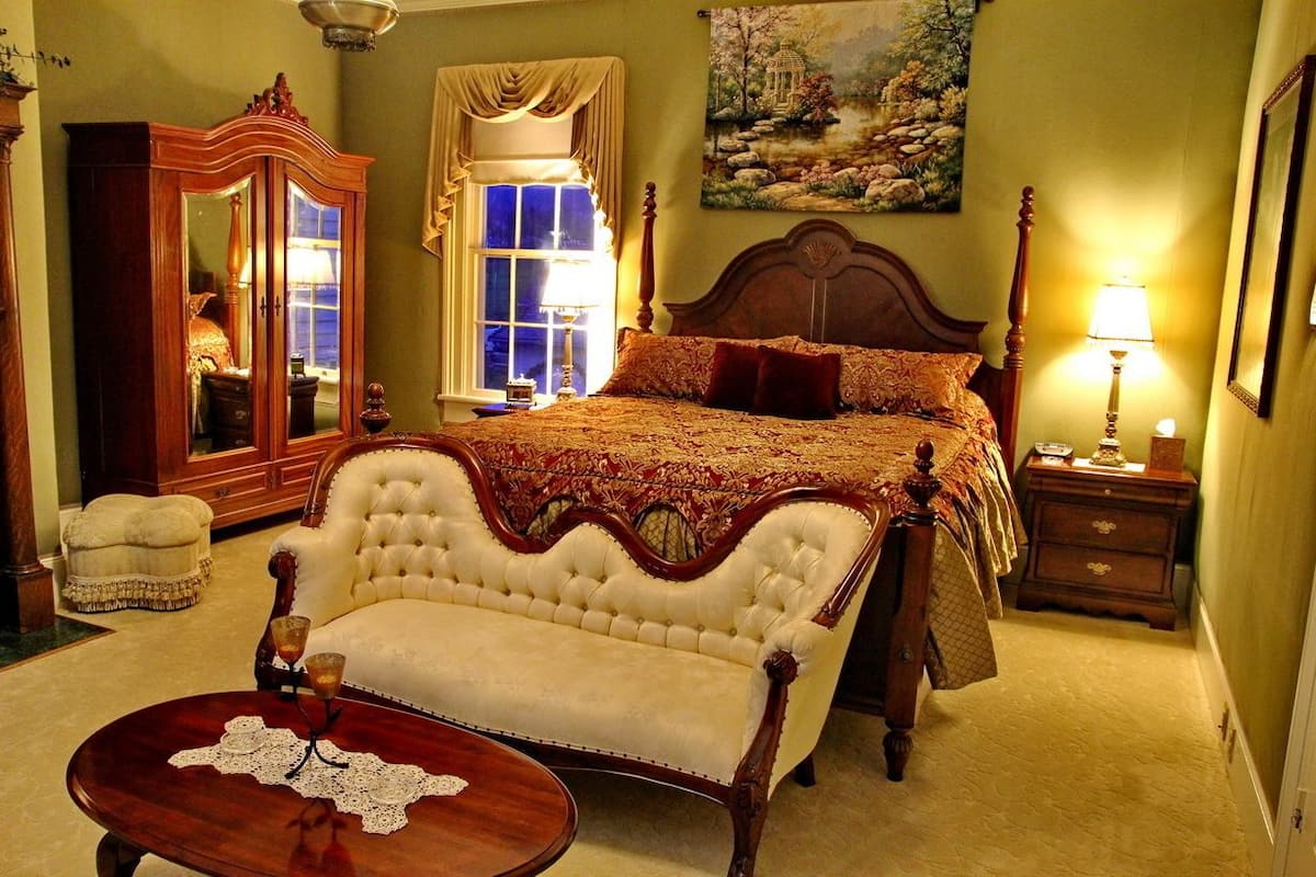Quarto com uma cama grande, um guarda-roupa de madeira, uma cadeira, uma mesa pequena com copos e um quadro na parede.