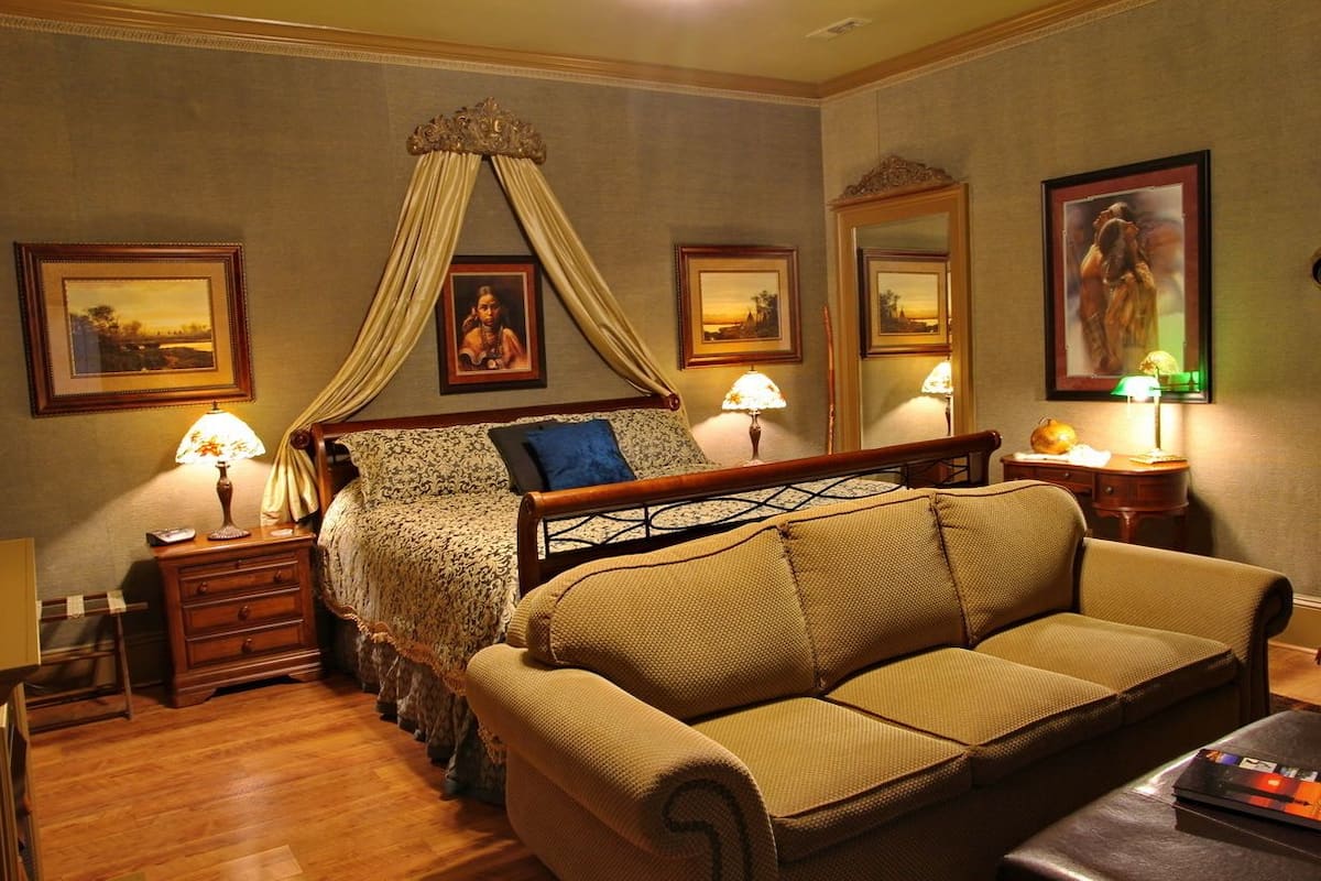 Quarto com cama de dossel, sofá, mesas de cabeceira com luminárias, quadros nas paredes e um espelho.