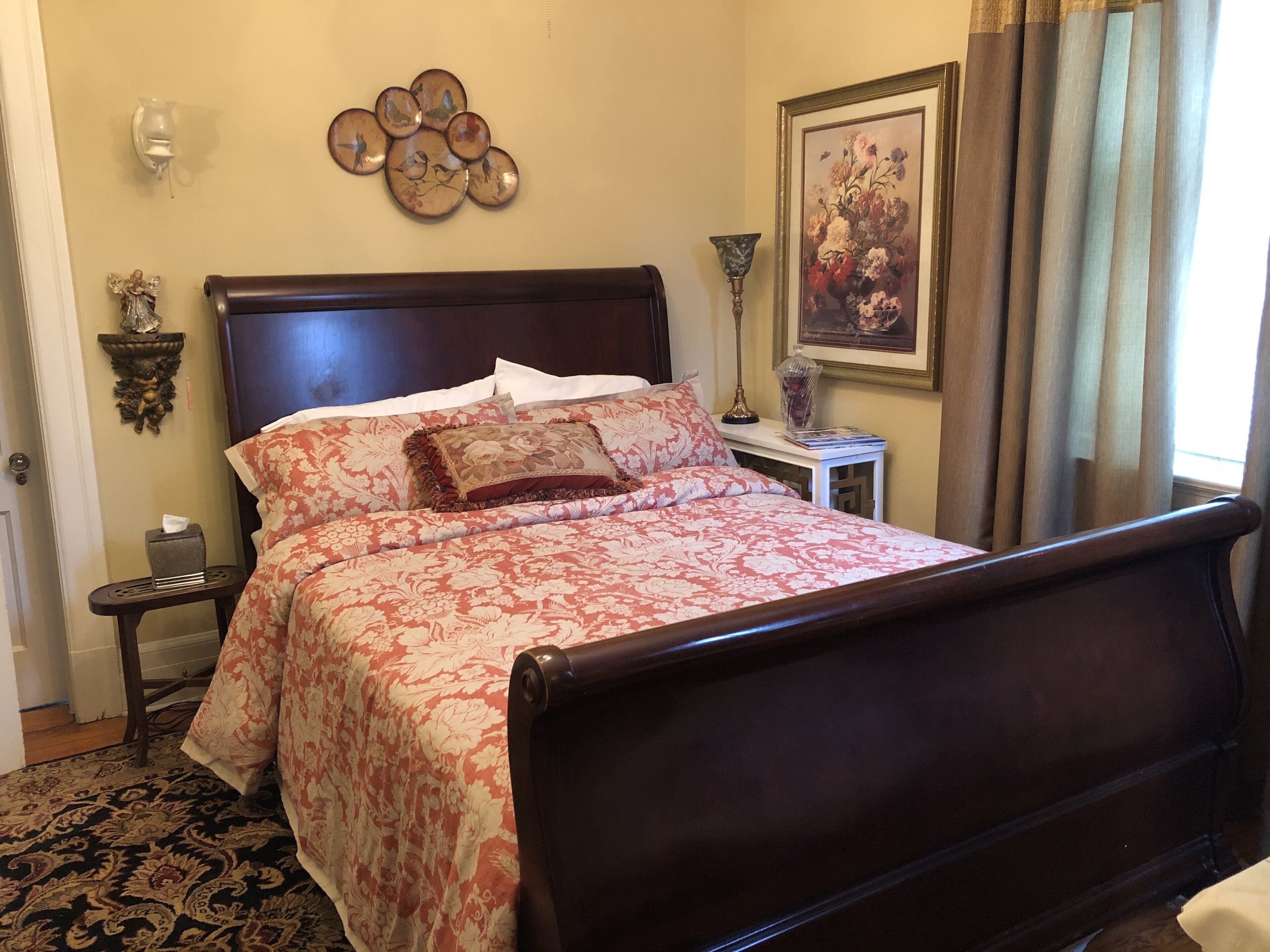 Una cama bien hecha con ropa de cama floral, un cabecero de madera, una mesita de noche con una lámpara y un adorno en la pared.