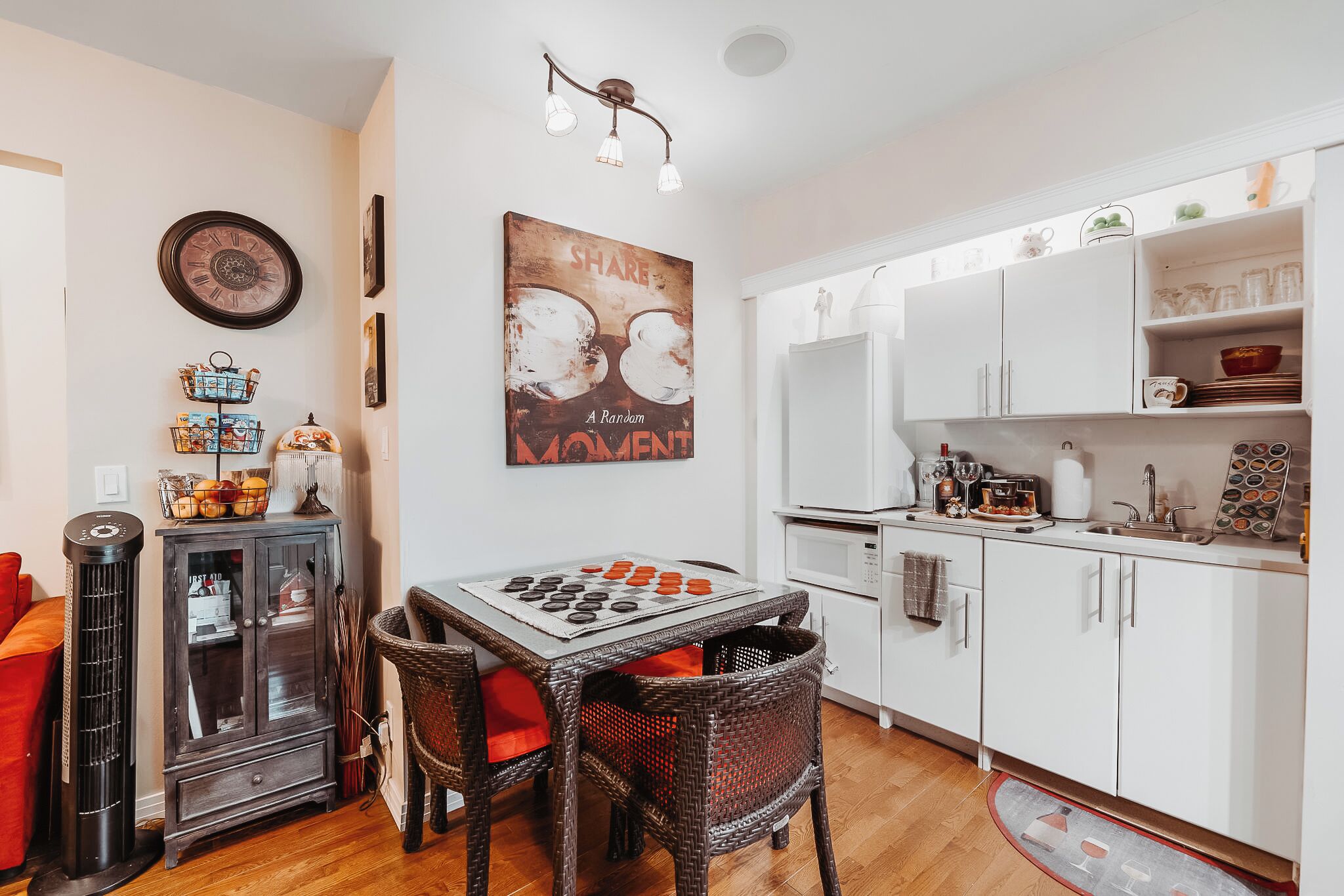 A compact kitchen with white cabinets, a dining table with chairs, a standing heater, and a wall-mounted clock.